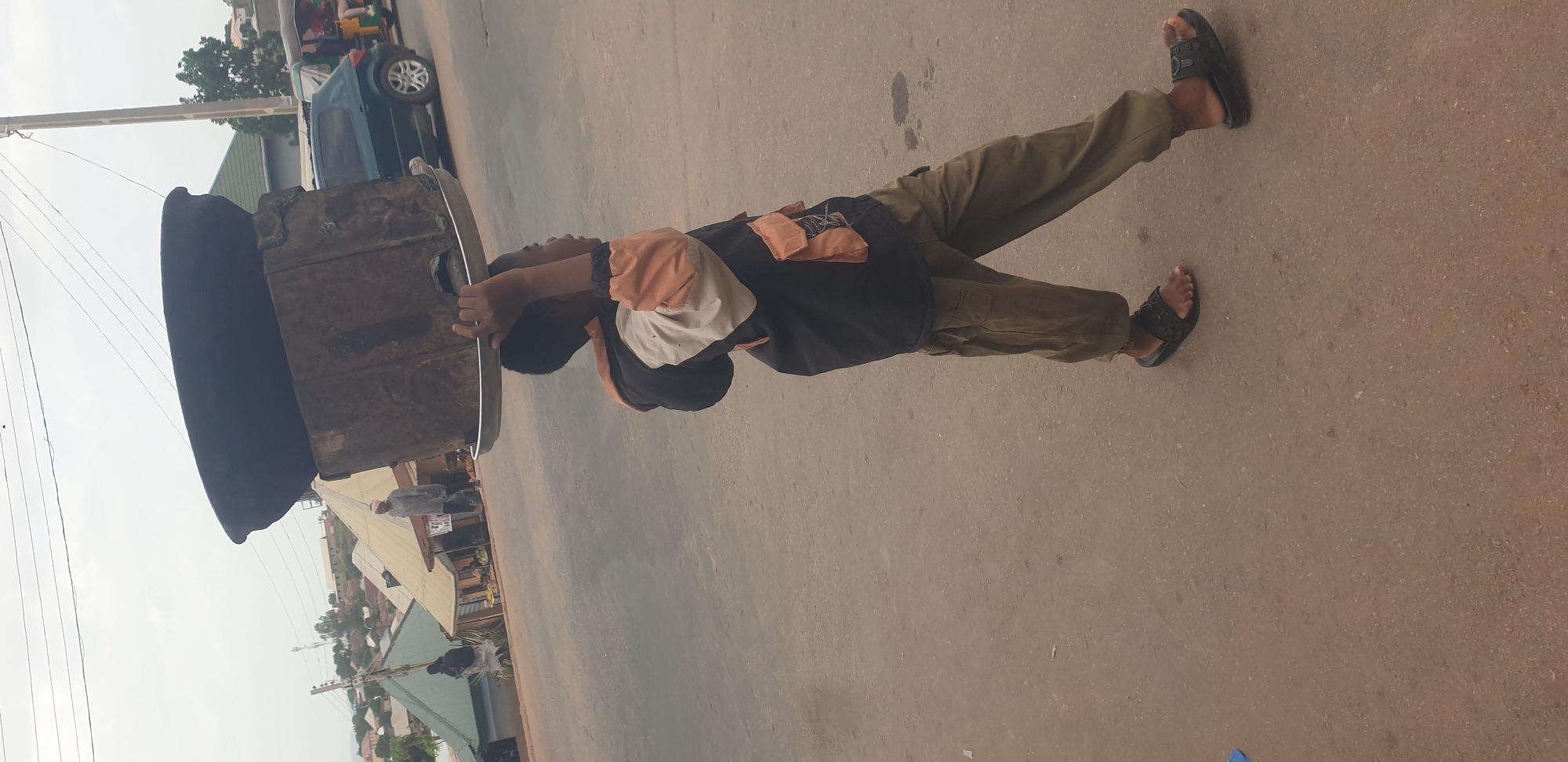 A young boy, dressed in a black and orange shirt and green pants, stands on a paved road in Barakin Lerawi, Nigeria. He is seen from the side, facing the right, and appears to be holding onto a large, dark, circular object, possibly a lid for a container. The object is partially obscuring his body. His feet are clad in black sandals.
The background reveals a street scene with buildings and a cloudy sky, suggesting it is daytime. A small blue vehicle and a white pole are also visible. The overall impression is a candid shot of a child engaged with an object on a typical Nigerian street. There is no visible text in the image.
