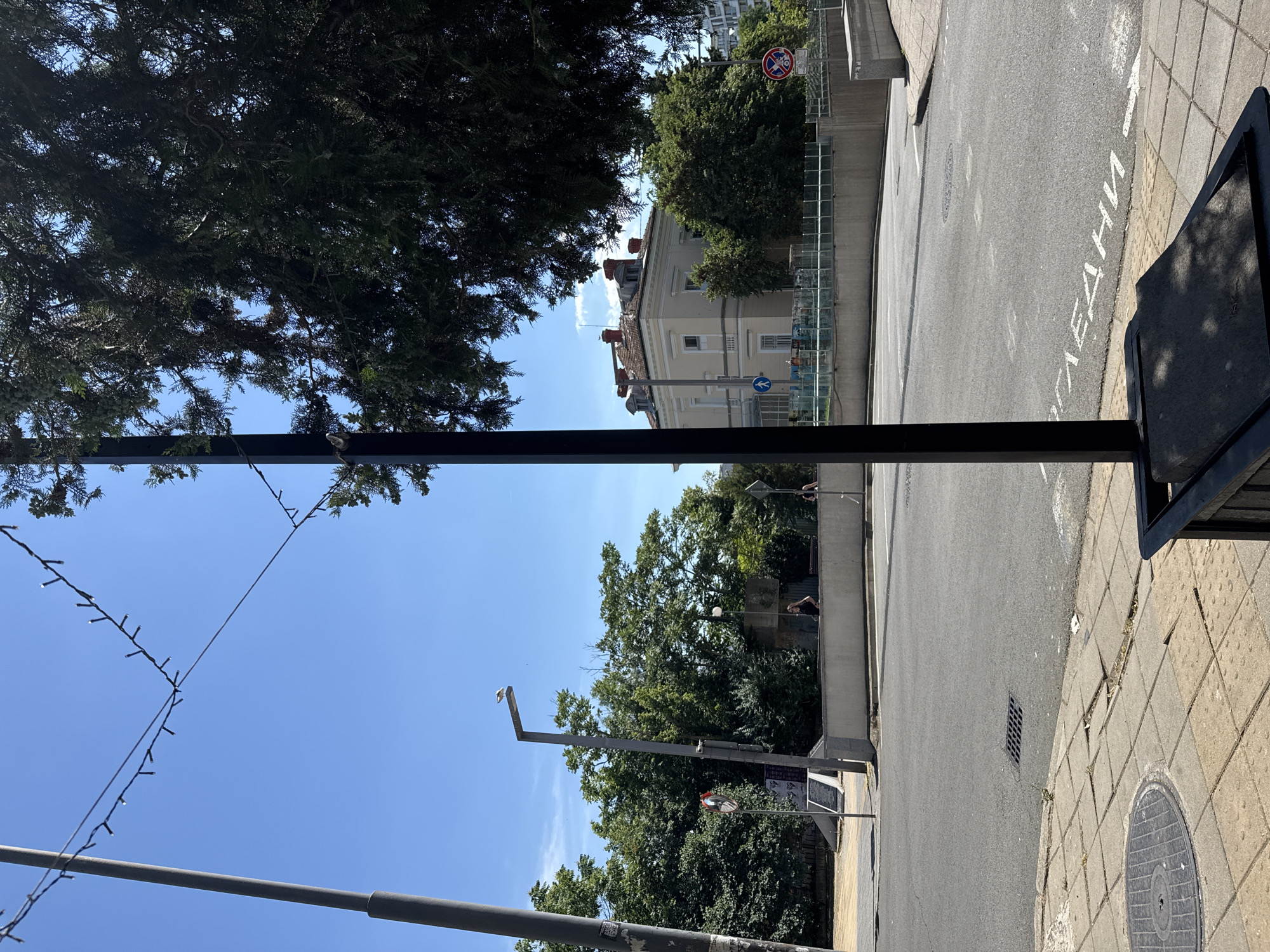 In Burgas, Bulgaria, on a clear, sunny day, a street scene unfolds with a focus on an intersection. A black lamppost stands prominently in the foreground, casting a shadow on the paved sidewalk. The sidewalk itself is made of square concrete tiles, some of which are cracked or uneven. A manhole cover is visible on the left side of the sidewalk.
Beyond the sidewalk, a two-lane asphalt road curves into the distance. White text, likely Cyrillic script, is painted on the road surface, pointing towards the right. A storm drain is present in the road.
In the middle ground, a light-colored, multi-story building with a red-tiled roof and dormer windows stands behind a low, concrete barrier. Trees, dense and green, surround the building and line the street. Several individuals are visible in the middle ground, including two people sitting on a bench behind the concrete barrier and another person standing near the building's entrance.
Further back, more trees and what appears to be another building can be seen. A traffic light pole with a sign depicting a pedestrian crossing is positioned on the left side of the road. A circular red and white traffic sign, indicating a prohibition, is visible on the right side of the road. A string of small lights is strung across the sky between poles, likely for decoration. A bird is perched on top of a lamppost on the left.
