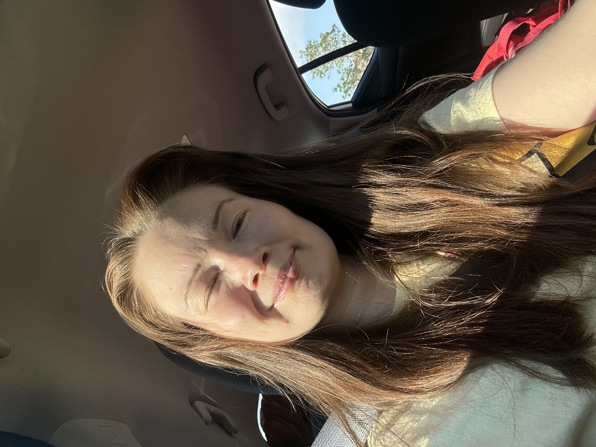 A woman is pictured in a car in Colorado Springs, Colorado. She has long, brown hair and is wearing a yellow shirt. Sunlight is shining on her face, causing her to squint. The interior of the car is visible, as is a window showing a tree and the sky. The image appears to be a selfie.