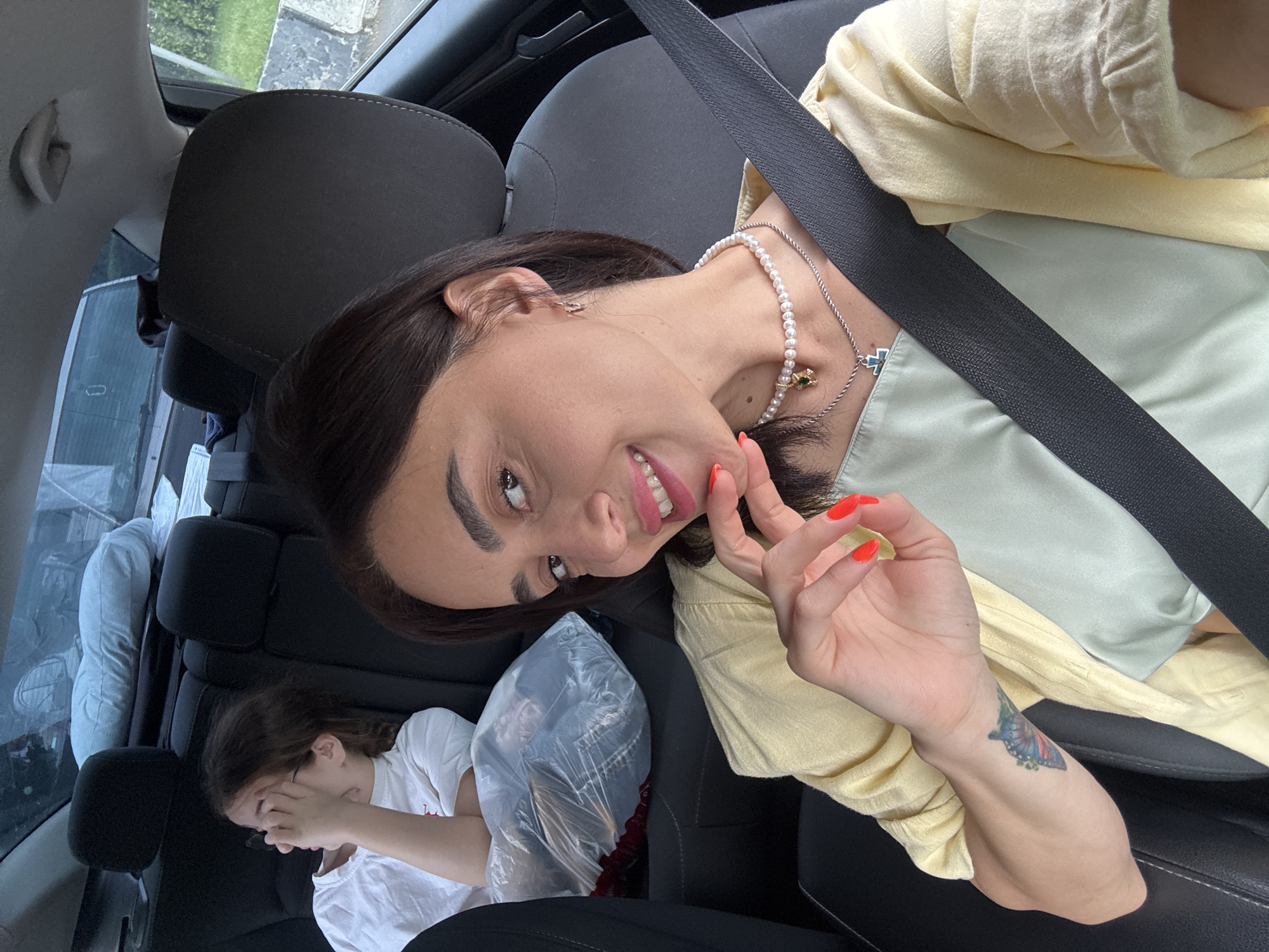 A woman with dark hair and bright pink lipstick smiles at the camera, her left hand touching her chin. She is wearing a light yellow cardigan over a pale green top and a pearl necklace. A seatbelt is visible across her chest. In the background, a young girl with her hand covering her eyes is seated in the backseat of a car, along with a large plastic-wrapped bag. The interior of the car appears to be dark. The image is taken from inside the car, looking forward.