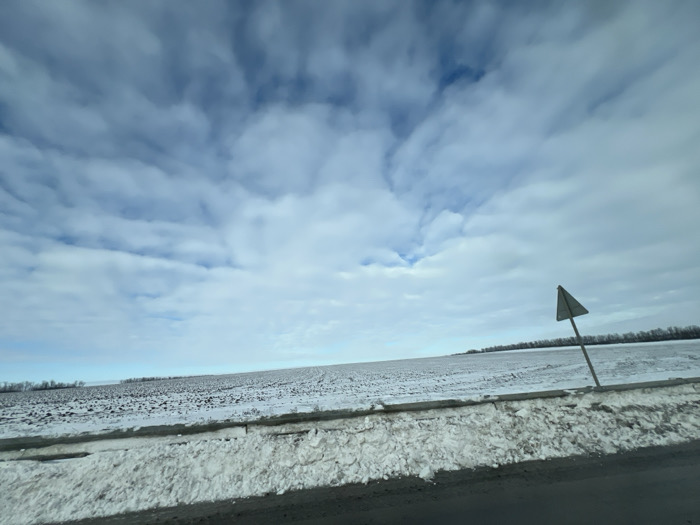 The image depicts a winter landscape in Khapry, Russia, characterized by a vast, snow-dusted agricultural field under a cloudy sky. The sky is predominantly light blue and white, filled with a dense layer of stratocumulus clouds, with occasional patches of deeper blue sky visible. A dark-colored road occupies the lower foreground, bordered by an irregular, substantial bank of white snow. Parallel to the road and partially obscured by the snow bank is a low, dark barrier or structure. On the right side of the frame, a light-colored, triangular sign mounted on a pole leans slightly left, positioned atop the snow bank near the road. The field, stretching to the horizon, displays uneven snow cover, revealing dark brown or black tilled earth in patches. A distant line of leafless trees or vegetation forms a dark band along the far horizon. No individuals or active subjects are present.