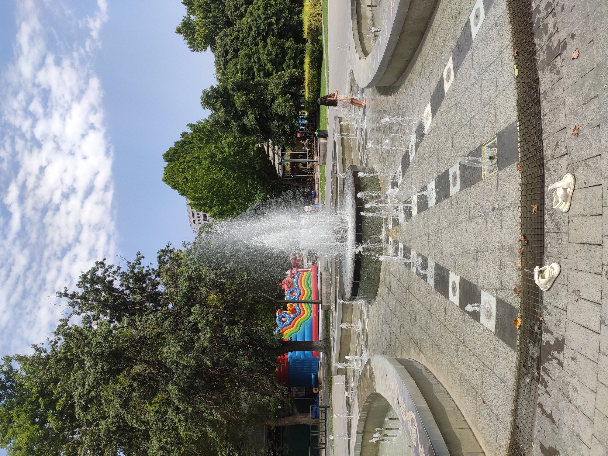 In an outdoor urban park, a large multi-tiered fountain is active with water jets spraying upwards. The fountain is surrounded by a paved area with a patterned tile design. In the foreground, on the paving stones, are two white sandals. To the left of the main fountain, a colorful inflatable bounce house with slides and characters is visible, suggesting a children's play area. In the background, lush green trees form a dense canopy, and a building with a modern facade can be seen peeking through the foliage. The sky above is a clear blue with scattered white clouds. A young girl in a light-colored dress is standing in the shallow water of the fountain, looking towards the right.
