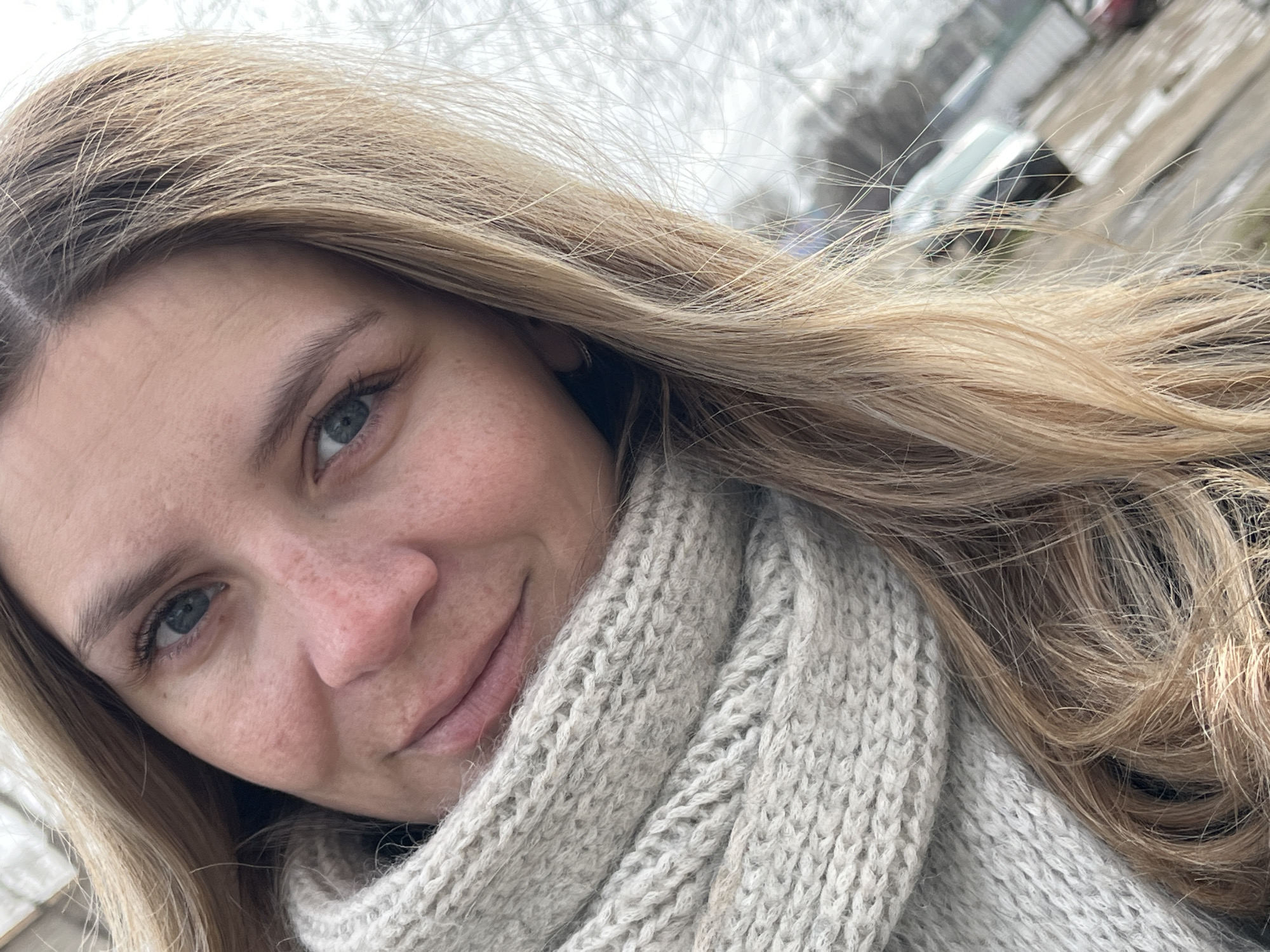A close-up, selfie-style photograph depicts a fair-skinned woman with light brown and blonde hair, partially obscuring the background. Her hair is long and flows to one side, partially covering a thick, light grey knitted scarf. She has blue eyes and visible freckles on her face. The background is slightly blurred, showing trees without leaves and a car parked in the distance. Based on the objects visible in the background, the photograph was likely taken outdoors. While a specific location in Chaikovsky, Russia, cannot be definitively ascertained from the image, it appears to be a neutral, urban environment. The subject is smiling, and there are no visible actions or interactions apart from the self-portrait.