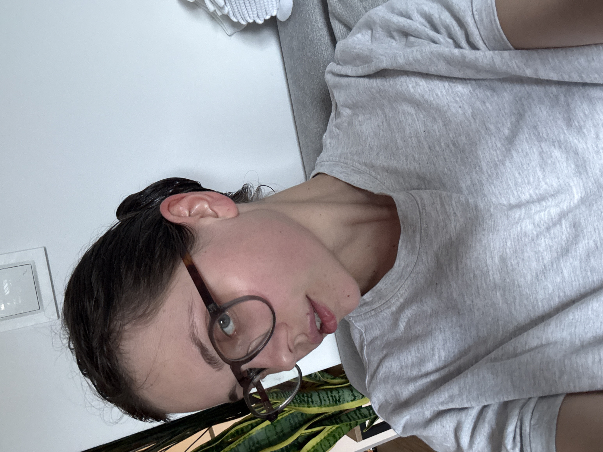 The media file shows a close-up, rotated portrait of a person wearing glasses and a light grey t-shirt. The person has dark hair pulled back and is looking off to the side. Their expression appears neutral. The background is predominantly white, with a hint of a plant with green and yellow striped leaves visible at the bottom. A light grey piece of furniture, possibly a sofa or chair, is also visible in the background. The lighting suggests an indoor setting. There is no visible text in the image. The location context provided is Szklarska Poręba, Poland, but this information cannot be visually confirmed from the image itself.