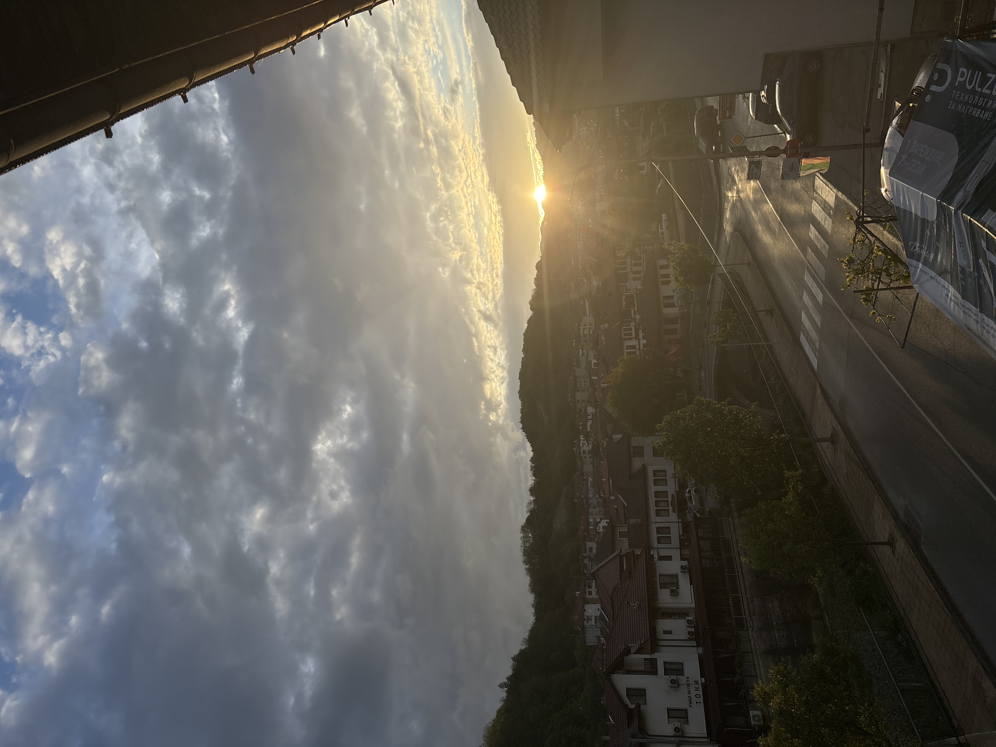 The image depicts an elevated view of a street in Zlatograd, Bulgaria, with the sun setting over a tree-covered hill in the distance. The sky is filled with cloudy skies, with a prominent building overhang in the upper left corner of the frame. A road runs vertically, with cars parked along one side and a zebra crossing visible. Buildings line the road, and an advertising billboard displaying the text "PULZE" is in the right side of the image.