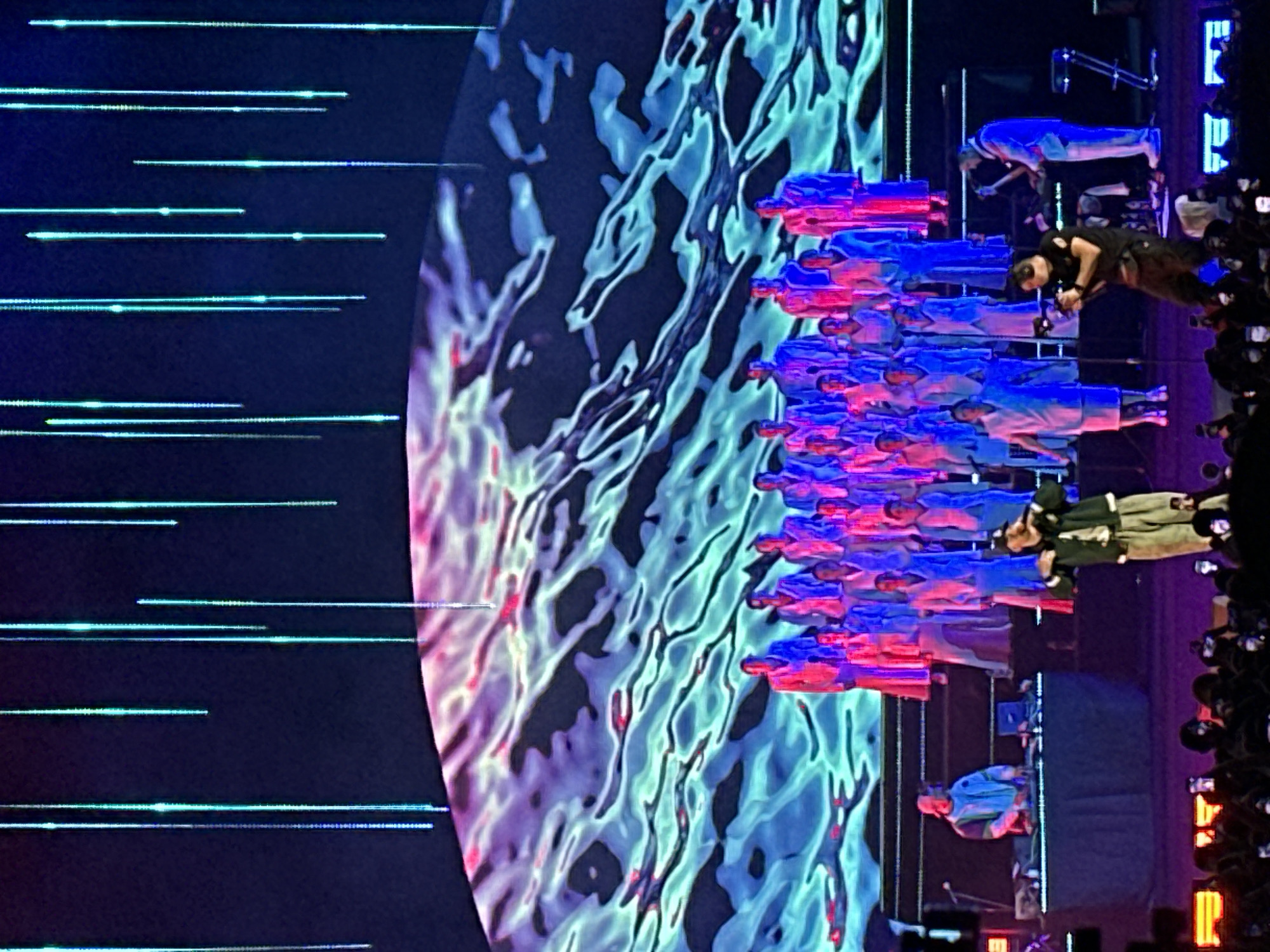 This is a medium shot, captured from a low angle, of a live musical performance. The background is dominated by a large screen displaying an abstract, wavy pattern of blues and purples, with vertical, thin blue lights descending from the top. In the middle ground, a choir of individuals, all dressed in matching light-colored outfits with brightly colored accents (red, blue, purple), are arranged in tiered rows on a platform.
In the foreground, on the left, a DJ is seen behind a table with equipment, wearing a light blue hooded jacket and a cap. To the right of the DJ, closer to the audience, a male performer in a dark jacket and light-colored pants is rapping or singing, with his hands in a gesture. Further to the right, another performer in a blue jacket and white pants is playing a keyboard, and a cameraman is operating a camera on a tripod. The stage is lit with purple and orange lights, reflecting on the floor. The audience in the very foreground is partially visible, with many people holding up phones to take pictures or videos. The overall scene suggests a large-scale concert or performance event. The image location is not determinable from the visual information provided.