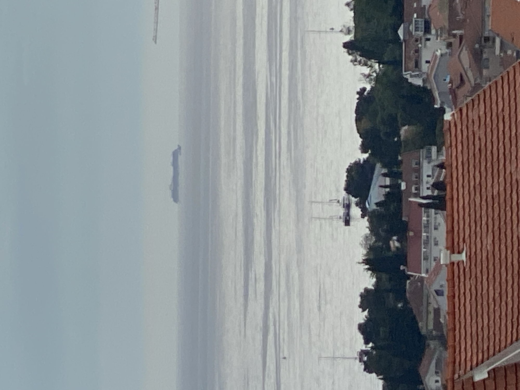 The image depicts a view from an elevated position, possibly a rooftop, overlooking the Bay of Budva in Montenegro. The calm water reflects the sky, creating a shimmering effect. A large ship is visible on the water, situated in the distance. Along the shoreline, there are buildings and trees suggesting a populated coastal area. The architectural style suggests a Mediterranean setting. The presence of tiled roofs in the foreground indicates residential construction. There are several masts in the water, indicating sailboats. The sky is overcast, and visibility is somewhat limited. The image captures the scenery of a coastal city, likely Budva.