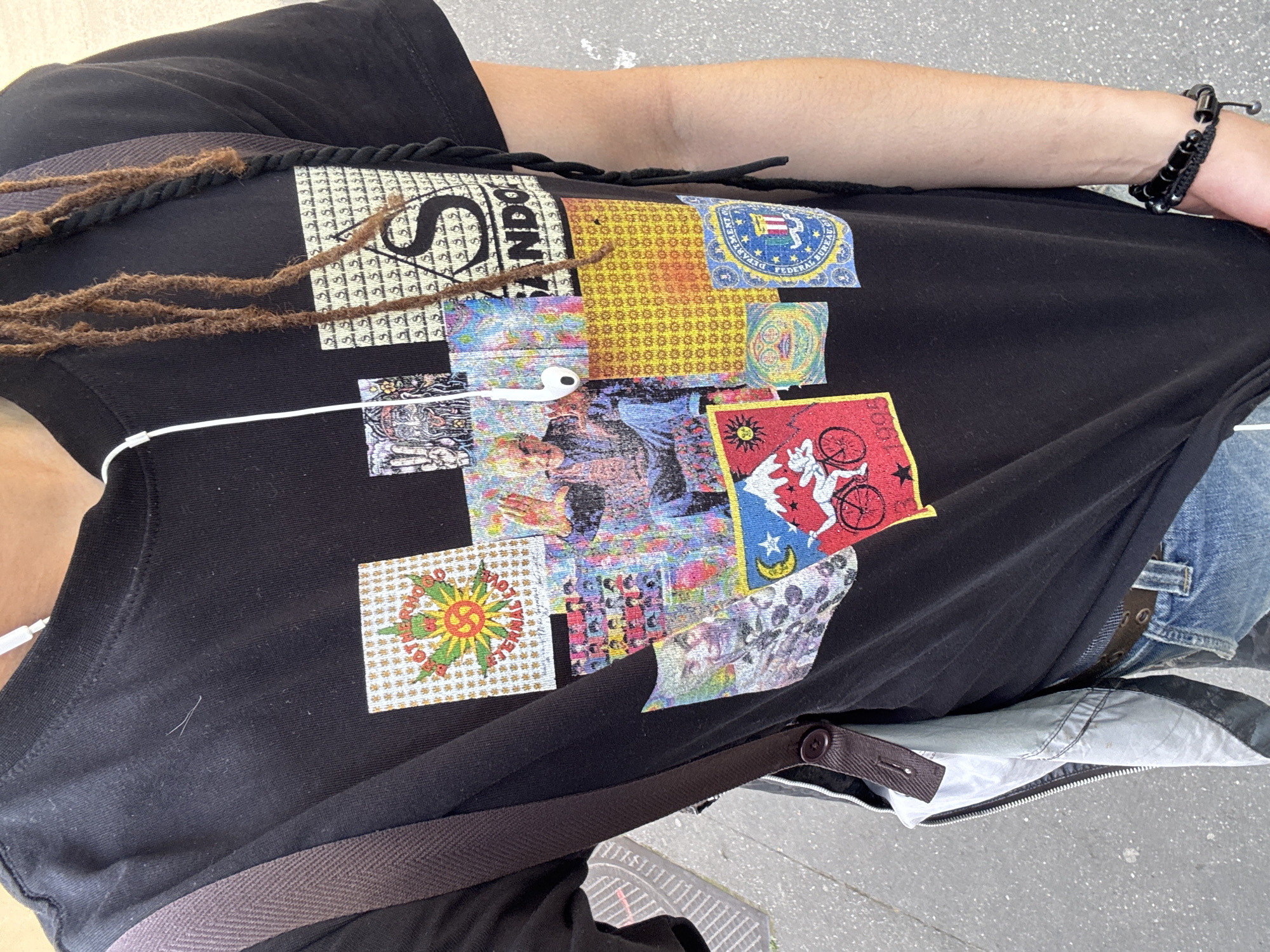 A person is wearing a black t-shirt with a collage of various images printed on the front. The images include what appears to be a logo, possibly a seal, and abstract patterns. The person has dreadlocks and is wearing a black bracelet on their right wrist. A white earbud wire is visible, leading to an earbud tucked under the shirt. They are also wearing a brown strap across their chest. The person appears to be outside on a paved surface. Part of a pair of blue jeans and a grey jacket are visible on the right side of the image. The location is unspecified; there is no way to know if this image was taken in Paris, France.