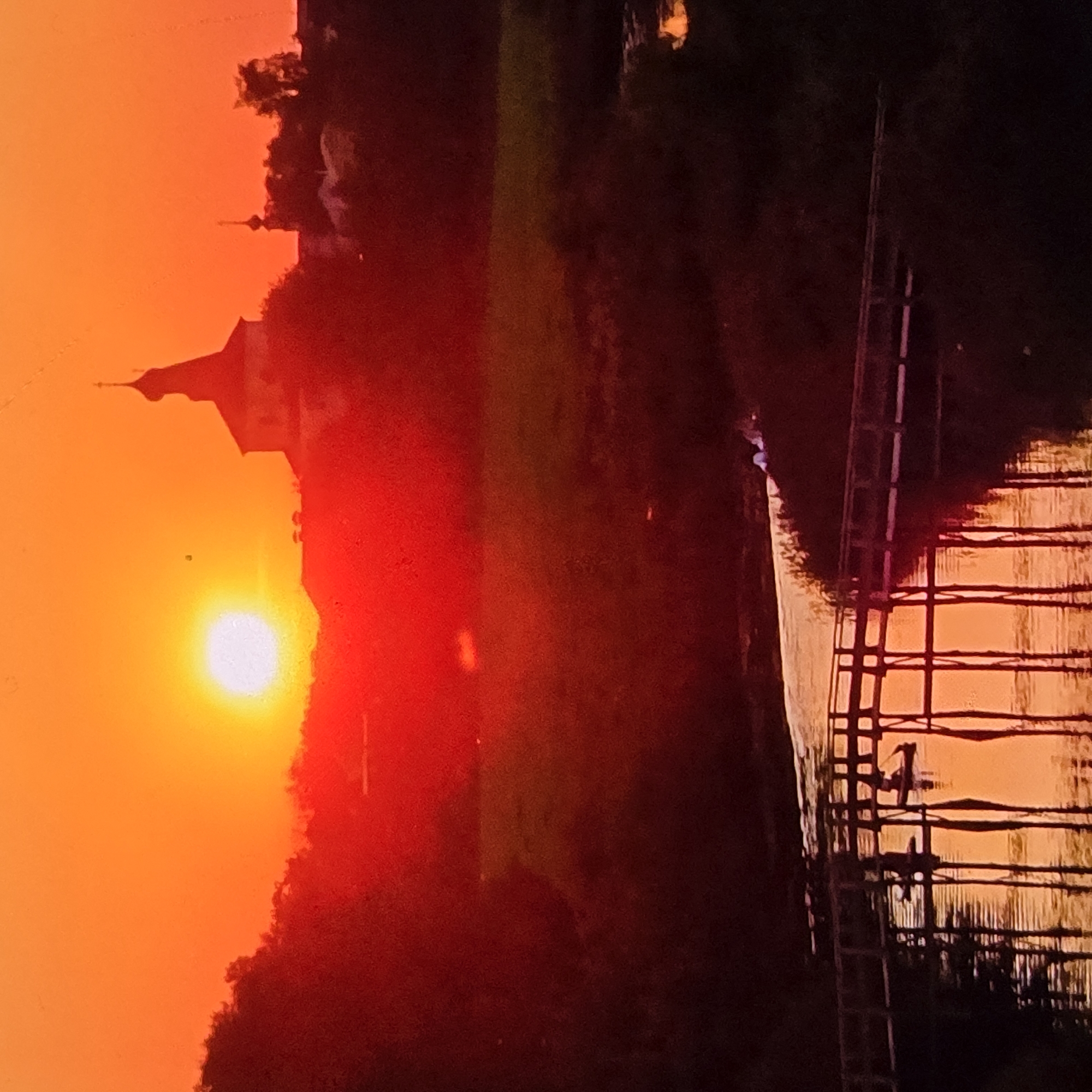 The image captures a vibrant sunset over a rural landscape, likely on the outskirts of Vladimir, Russia. The sky is ablaze with fiery orange and red hues, with the sun a bright, incandescent disc near the horizon. Silhouetted against this dramatic backdrop are the distinct shapes of church architecture, characterized by domed roofs and steeples, characteristic of Russian Orthodox churches. One prominent church dominates the middle ground, its structure clearly outlined.
In the foreground, a wooden footbridge spans a tranquil river. The bridge is supported by numerous vertical pilings that cast long, dark reflections on the water's surface, mirroring the warm tones of the sunset. The river itself appears calm, its surface reflecting the sky and the bridge. On or near the bridge, there are faint indications of human presence. Small, dark shapes suggest figures or possibly a boat on the water near the bridge, and in the upper right portion of the frame, there appear to be a few scattered lights or small structures on land, indicating a settlement or scattered houses. The overall scene evokes a sense of peacefulness and natural beauty, enhanced by the dramatic lighting of the sunset.