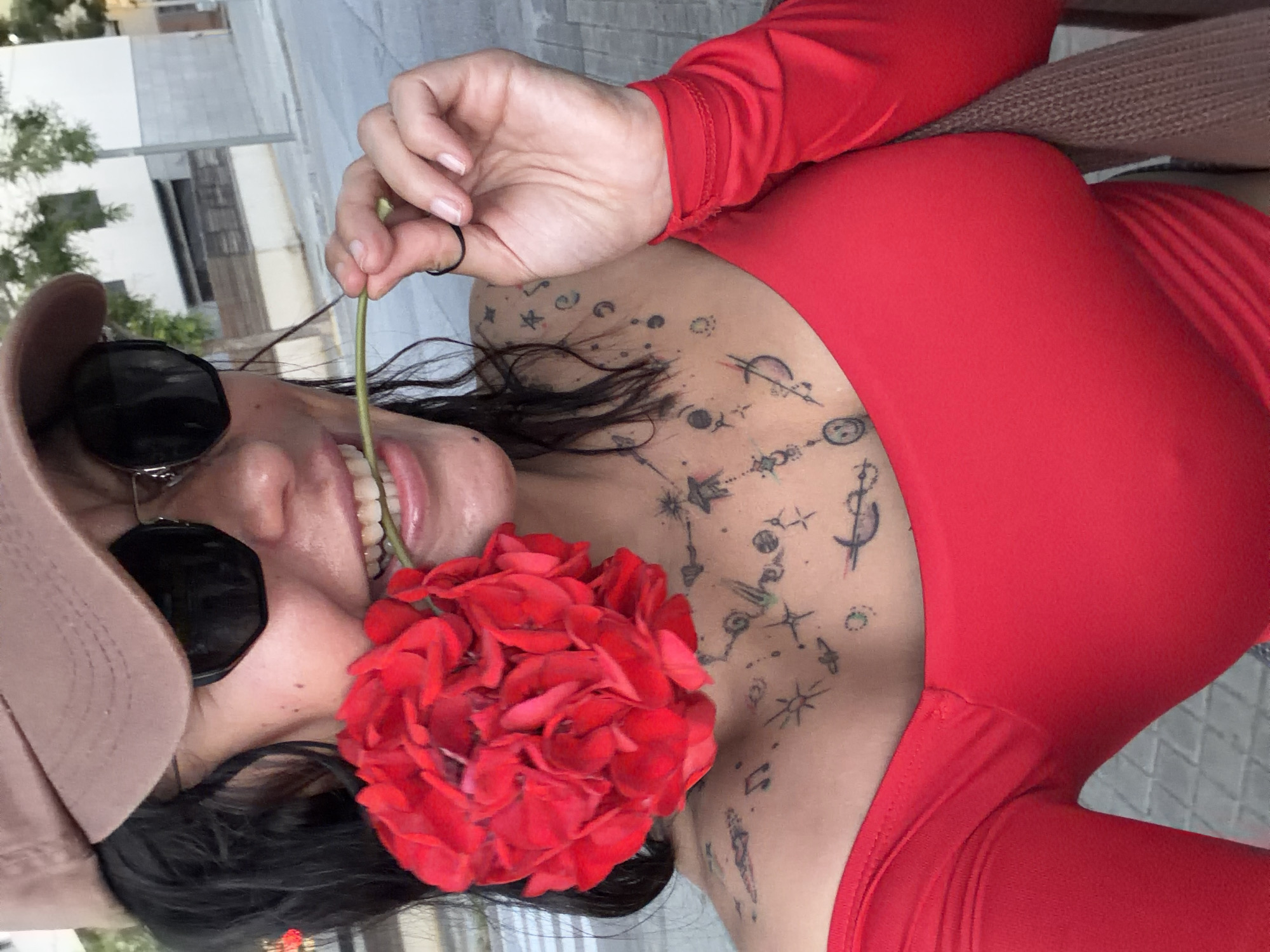 A woman with dark hair is seen up-close holding a red flower in her mouth. She is wearing sunglasses and a tan baseball cap. She also has a red long-sleeved top. She has numerous tattoos on her chest and upper body. The location appears to be an outdoor public space, possibly in L'Hospitalet De Llobregat, Spain, indicated by architectural details and trees in the background. She is sitting on a wicker couch.