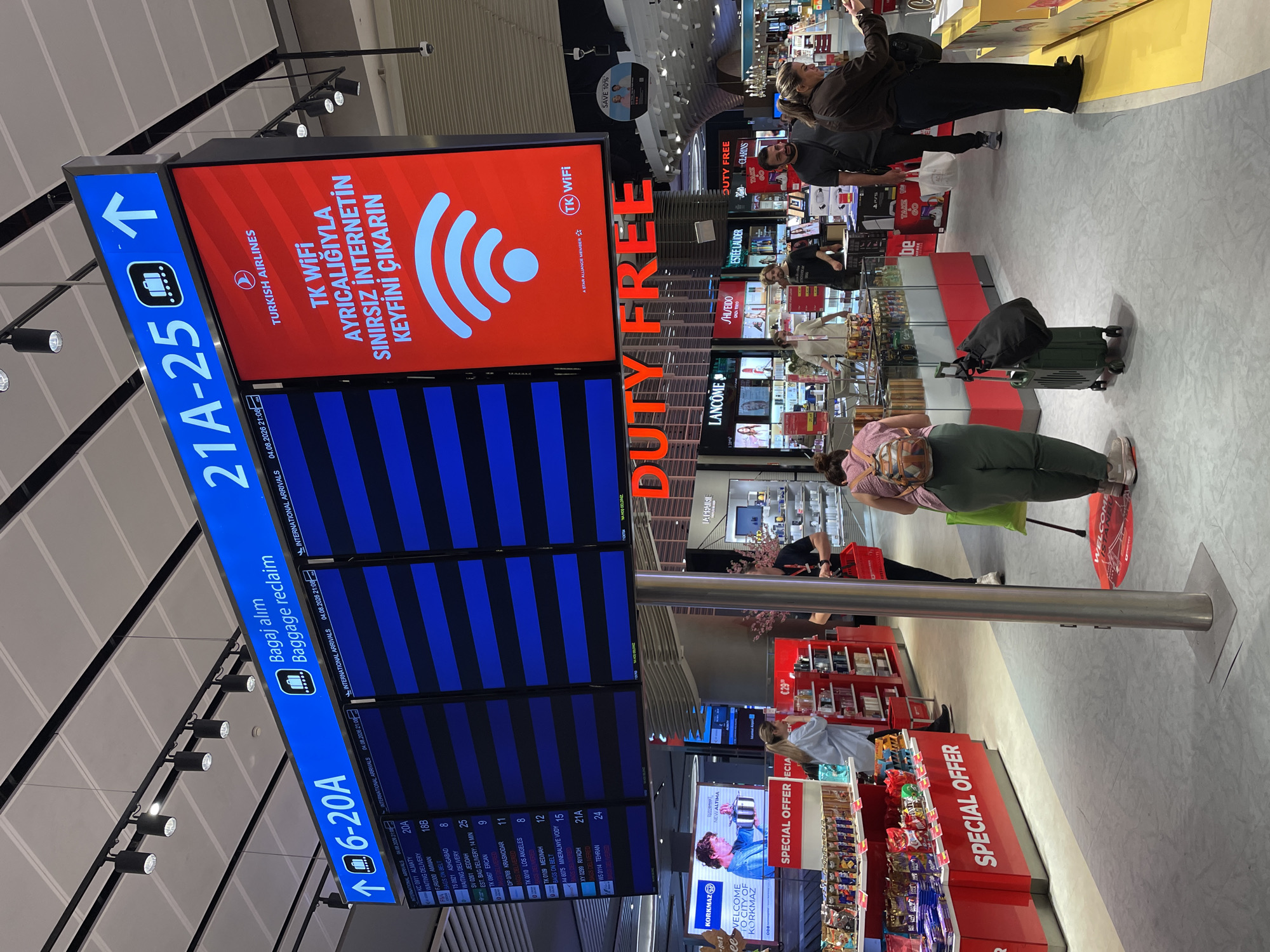 The image shows an interior view of the Arnavutköy, Turkey airport. In the foreground, there is a large digital display showing flight information. Above the flight information, there is a smaller sign that reads "Bagaj alım Baggage reclaim" and an arrow indicating a direction. To the right of the flight information display is a red screen with the Turkish Airlines logo and text in Turkish and English, advertising free Wi-Fi. Below the digital displays, there is a Duty Free shop with various products on display. Several people are seen walking through the airport, some carrying luggage. The environment appears to be clean and modern, typical of an international airport.
