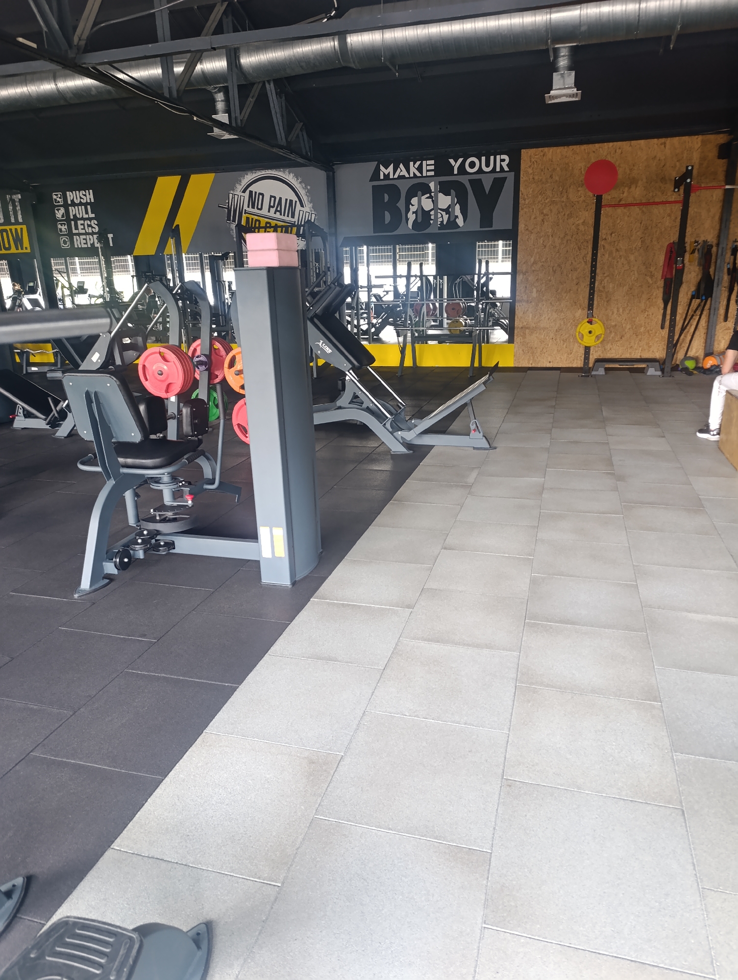 The image shows the interior of a gym, specifically the Zlatopil Sports Club, located in Zlatopil, Ukraine. The gym features various weightlifting equipment, including a leg press machine in the foreground, weight plates, and racks. The walls are adorned with motivational gym slogans, such as "PUSH. PULL. LEGS. REPEAT." and "MAKE YOUR BODY." The floor is covered with black rubber mats in one section and light grey tiled flooring in another. The overall environment suggests a functional and well-equipped fitness facility.