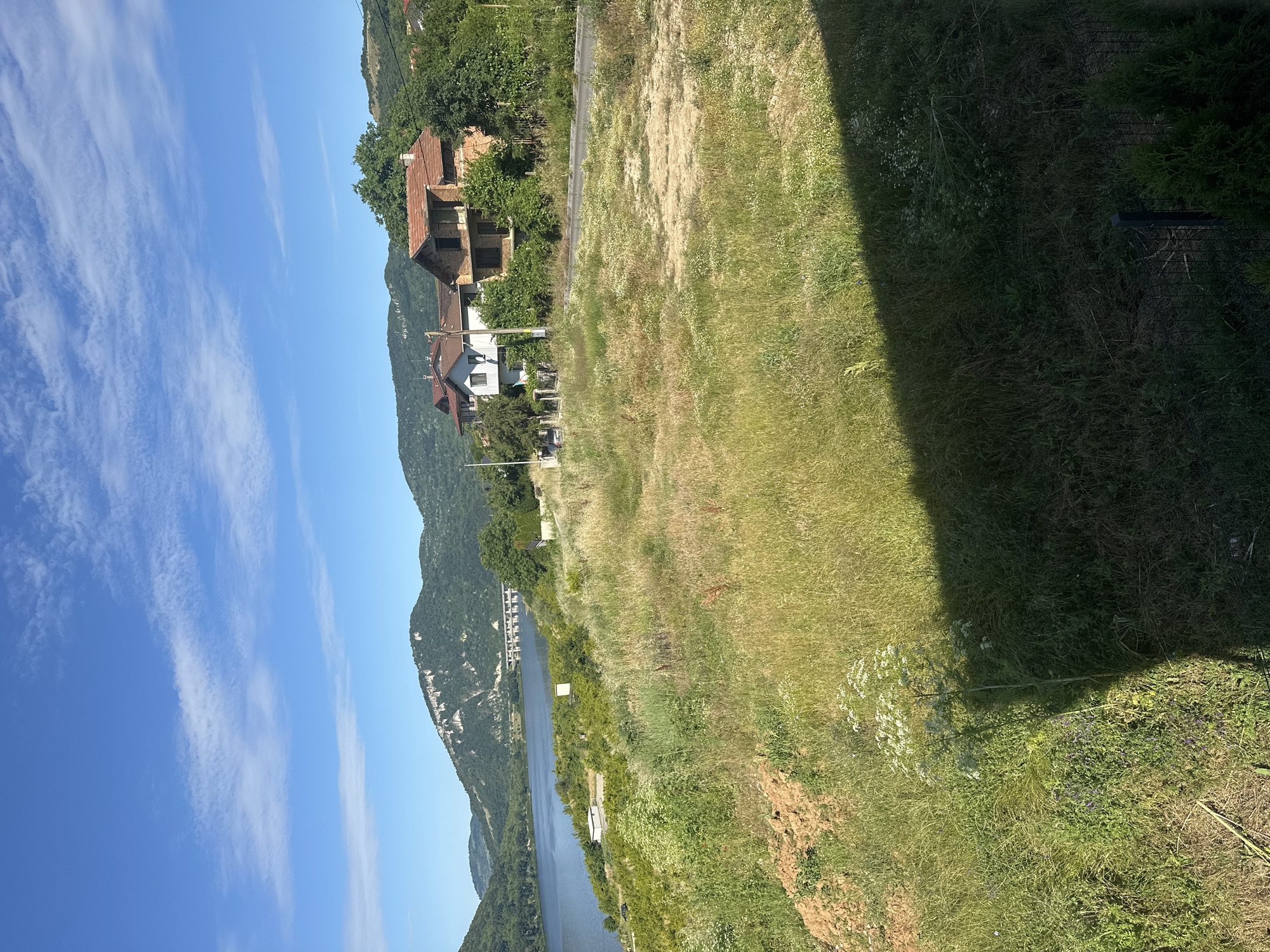 The image depicts a pastoral landscape under a clear blue sky with wispy clouds. A wide, calm body of water occupies the left side of the frame, with rolling green hills in the background. Several houses with red-tiled roofs are nestled on the slopes among trees, suggesting a small village or rural settlement. The foreground is a grassy field with scattered wildflowers and some dry patches, indicating a sunny day. A shadow from an unseen object falls across the right side of the foreground. The scene conveys a sense of tranquility and natural beauty.