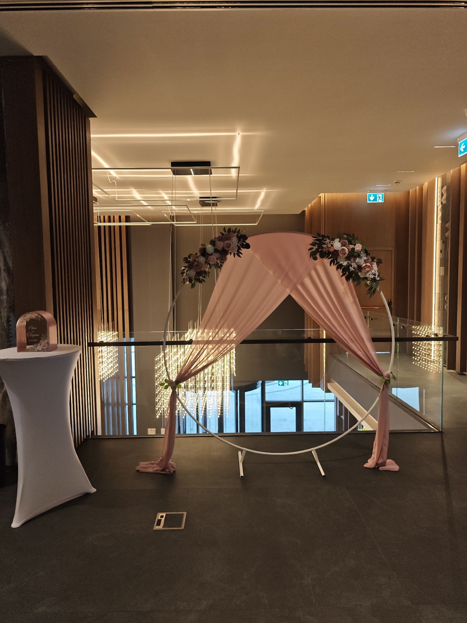 The image shows an indoor event space, possibly a wedding venue, in Sofia, Bulgaria. A circular white arch decorated with pink flowers and draped with pink fabric stands prominently. To the left, a white-covered table holds a framed sign. The floor is dark and tiled. Background elements include wood-paneled walls, glass railings, and modern lighting fixtures. The location appears to be on an elevated platform or mezzanine overlooking another level with windows. Exit signs are visible.