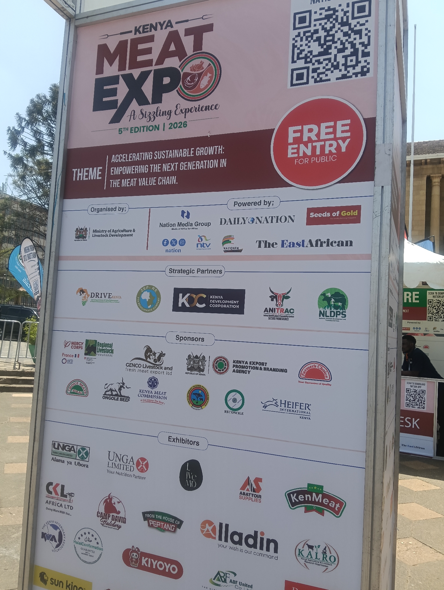 This is a full-frame, eye-level, medium shot of a large, pink and maroon billboard advertising the "Kenya Meat Expo 2026". The billboard is located outdoors, possibly in a public square or exhibition area in Nairobi, Kenya, given the context of the advertisement. The top section prominently features the event's title, "KENYA MEAT EXPO", with the subtitle "A Sizzling Experience" and details about the "5TH EDITION | 2026". Below this, a large QR code is visible on the right side.
The central section of the billboard is dedicated to the theme "ACCELERATING SUSTAINABLE GROWTH: EMPOWERING THE NEXT GENERATION IN THE MEAT VALUE CHAIN." It also includes sections for "Organised by:", "Powered by:", and "Strategic Partners", listing various organizations and companies such as the Ministry of Agriculture & Livestock Development, Nation Media Group, Daily Nation, The EastAfrican, and others, accompanied by their logos. A prominent red circle with white text "FREE ENTRY FOR PUBLIC" is located on the right side.
The lower sections are labeled "Sponsors" and "Exhibitors", featuring a multitude of logos of businesses and organizations involved in the meat industry. Notable sponsor logos include GENCO Livestock and fresh meat export ltd, KENYA MEAT COMMISSION, and various government and private entities. Exhibitor logos include UNGA LIMITED, KenMeats, KALIRO, and others.
In the background, to the right of the billboard, a portion of a white tent structure and part of a classical-style building with columns is visible, suggesting an event venue. A man in a dark shirt is partially visible standing behind a table to the right of the billboard, near another smaller advertisement for the "Nation App". The sky is overcast, with some trees visible on the far left of the image. The overall scene depicts an outdoor exhibition or event setup with a focus on the meat industry in Kenya.