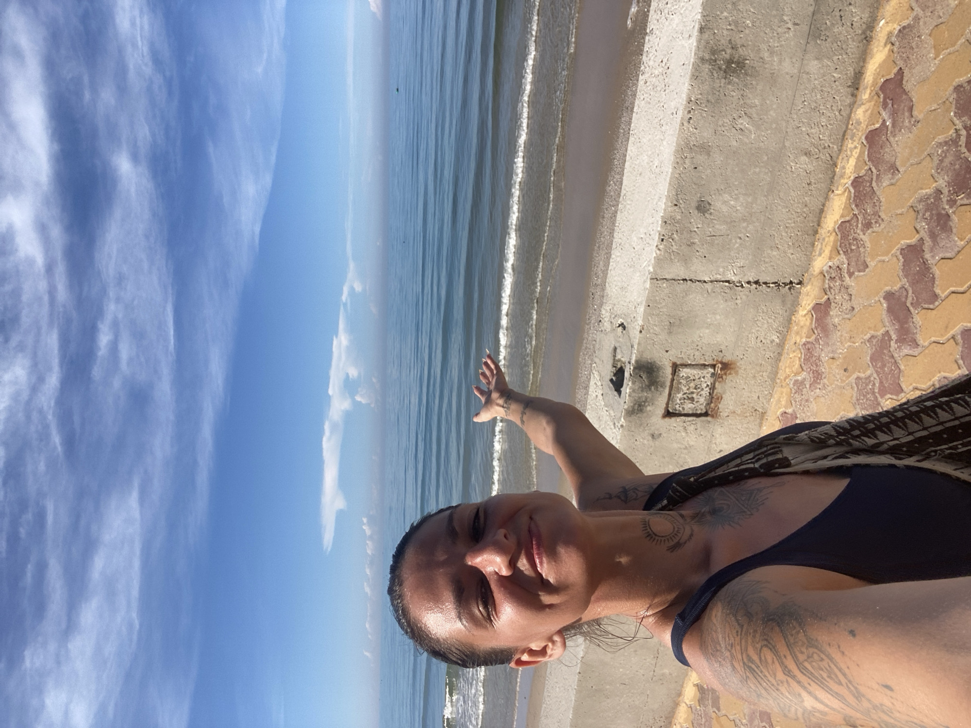 A woman with dark hair pulled back and tattoos on her arm and neck is taking a selfie on a sunny day. She is standing on a paved walkway next to a concrete barrier that overlooks the sea. The sea is calm with gentle waves, and the sky is blue with scattered white clouds. The woman is wearing a dark blue swimsuit and a patterned scarf draped over her shoulder. Her right arm is extended, palm facing outward, as if gesturing towards the ocean. The ground beneath her is paved with interlocking tan and reddish-brown tiles. In the distance, small boats can be seen on the water. The location appears to be a coastal area in Phu Thuy Ward, Vietnam.