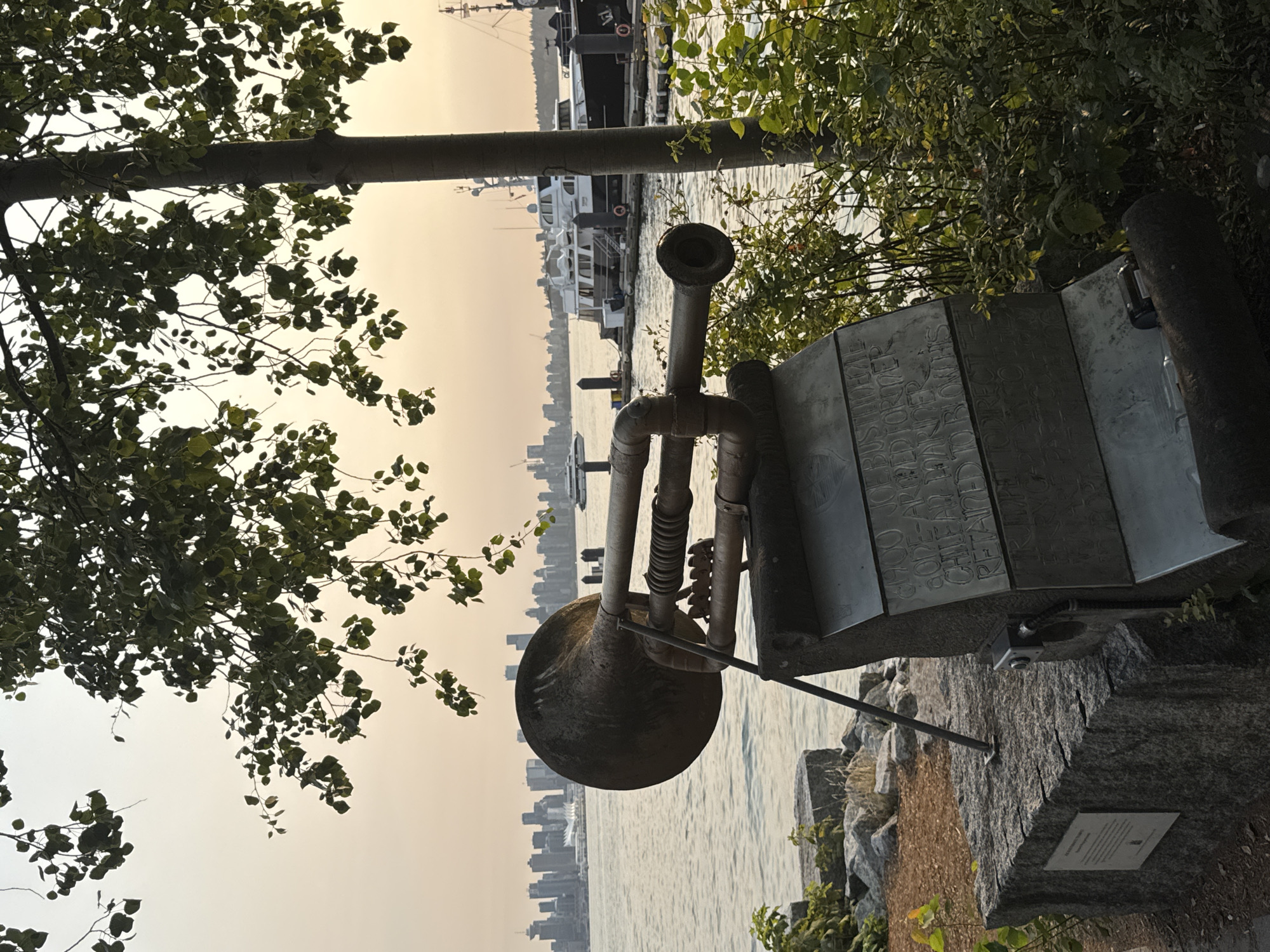 The image depicts an outdoor scene with a large, weathered brass horn sculpture in the foreground, mounted on a stone pedestal inscribed with text. The sculpture is positioned near the water, with a cityscape visible in the background across the body of water, which appears to be a harbor or bay. Several boats, including yachts, are docked in the water. Lush green foliage surrounds the sculpture and the base. The sky is overcast with a hazy, warm glow, suggesting either dawn or dusk. The sculpture's design is industrial and somewhat abstract, with visible pipes and a large bell. The stone pedestal has an information plaque attached to its left side and a small black camera mounted on its front. The overall impression is one of a public art installation in a waterfront urban environment. The cityscape in the background is likely Vancouver, Canada, given the prominent skyline features. The presence of a camera on the sculpture suggests it might be a functional installation, perhaps a sound sculpture or part of an interactive exhibit, and its placement in a public park or waterfront area indicates it is likely located in North Vancouver, Canada, a city known for its scenic waterfront views and public art.