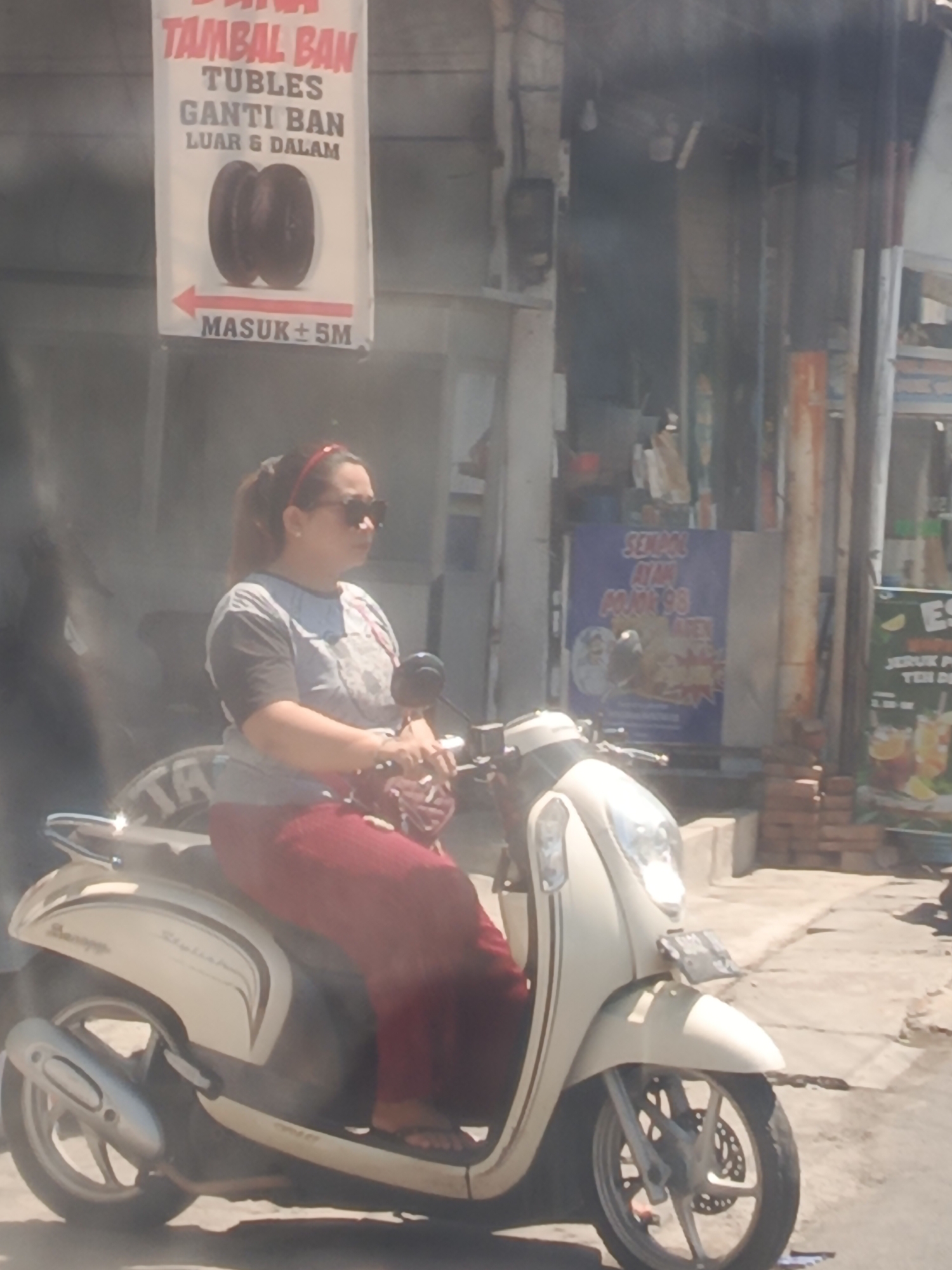 A woman is riding a white scooter in Sedati Agung, Indonesia. The woman is wearing a grey shirt with black sleeves, a red headband, and dark sunglasses. She is also wearing red pants and dark sandals. The scooter is a light cream color with silver accents and has a license plate that is difficult to read. The woman is looking to her right. In the background, there is a sign that says "TAMBAL BAN TUBLER GANTI BAN LUAR & DALAM MASUK ± 5M" which translates to "TUBE TIRE PATCH REPLACE TIRE INSIDE & OUTSIDE ENTER ± 5M". There are also other signs and buildings visible in the background, suggesting a commercial area. The scene appears to be a street in daylight.