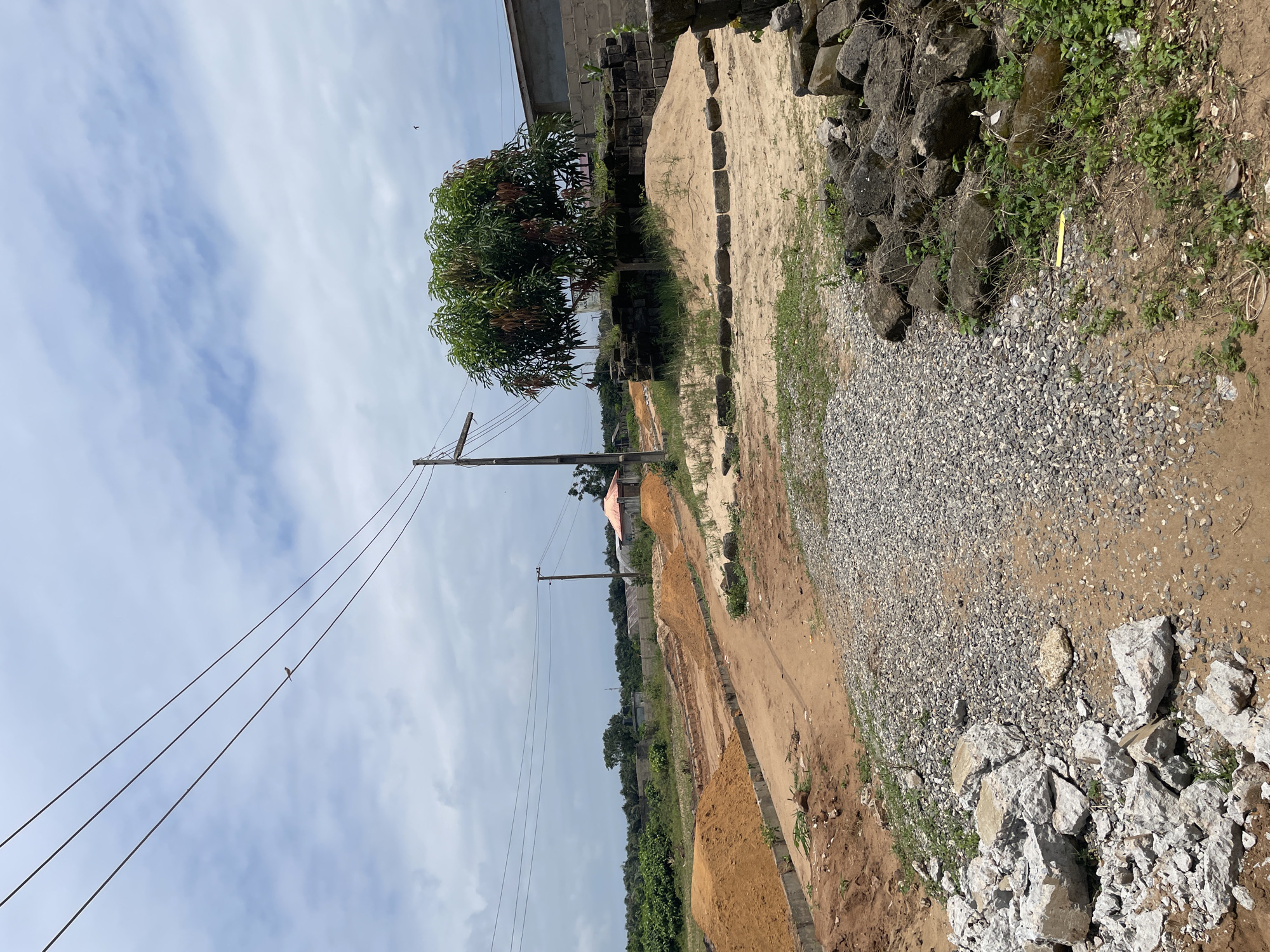 This is a photograph taken from a low angle, capturing a construction site in the city of Iyafin, Nigeria. The scene is dominated by a dirt road and piles of sand and gravel, indicative of ongoing construction work. Power lines stretch across the cloudy sky, supported by utility poles. To the right of the frame, a large mango tree stands amidst piles of bricks and sand. In the background, a small building with a red roof is visible, as well as other structures partially obscured by vegetation. The overall impression is one of a developing area undergoing significant transformation.
