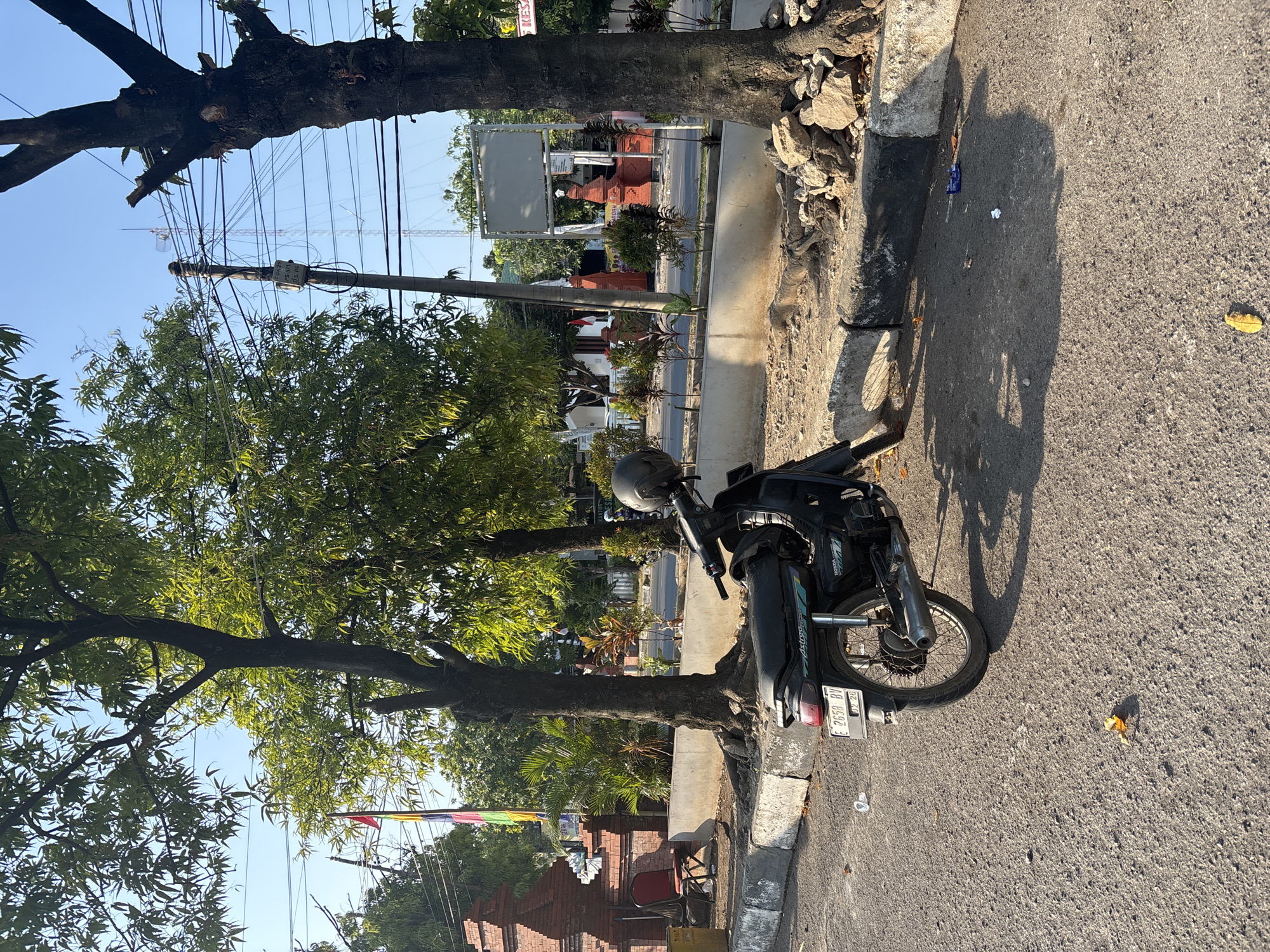The image shows a black Honda motorcycle parked on the side of a road in Cirebon, Indonesia. The motorcycle has a helmet placed on its seat. The road is paved and has a concrete curb. In the background, there are trees, a brick wall, a red chair, and some buildings. A tall telecommunications tower is visible in the distance. The sky is clear and blue. The scene appears to be during the daytime.