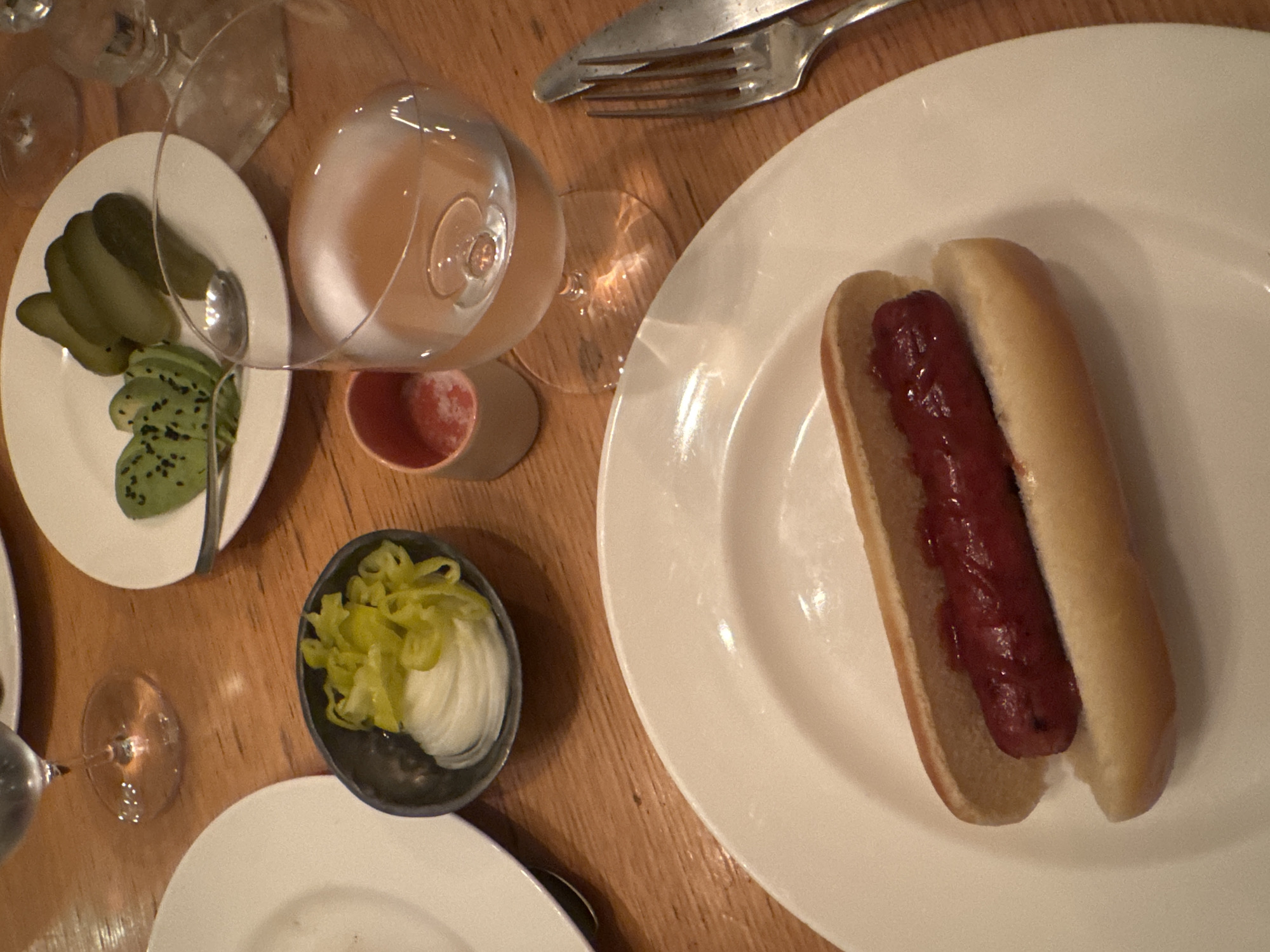 A high-angle shot captures a wooden table setting with various food items and utensils. A hot dog in a plain bun sits on a white plate, positioned on the right side of the frame. Adjacent to it, a fork and knife rest on the table. To the left, there's a plate containing sliced pickles and avocado with black seeds. A small bowl of shredded green peppers and onions is also present. A wine glass filled with a light-colored liquid and a small cup are placed near the pickle plate. Another plate and the base of a wine glass are partially visible in the lower-left corner. The scene appears to be set indoors, possibly in a restaurant or dining establishment. The location is not identifiable from this image alone; the city of New York is not discernible without additional information.