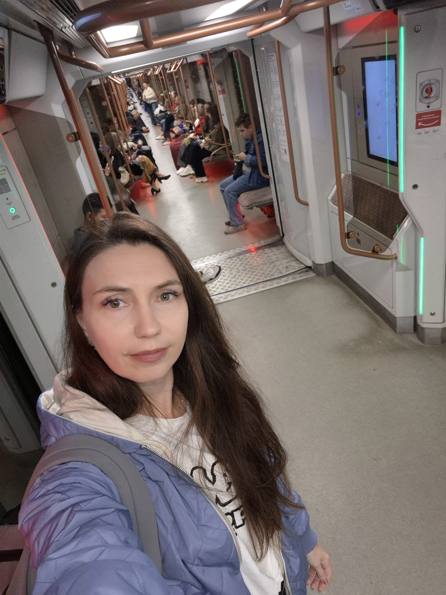 A woman with long brown hair takes a selfie in a train car. She is wearing a light blue quilted jacket over a white t-shirt and a grey backpack. The train car is modern with beige walls, copper-colored handrails, and fluorescent lighting. Several other passengers are seated in the train car, some looking at their phones. The floor of the train car is a light grey concrete. The woman's face is well-lit, showing her features clearly. The background is slightly blurred, emphasizing the woman as the main subject. The image appears to be taken inside a Moscow Metro train.