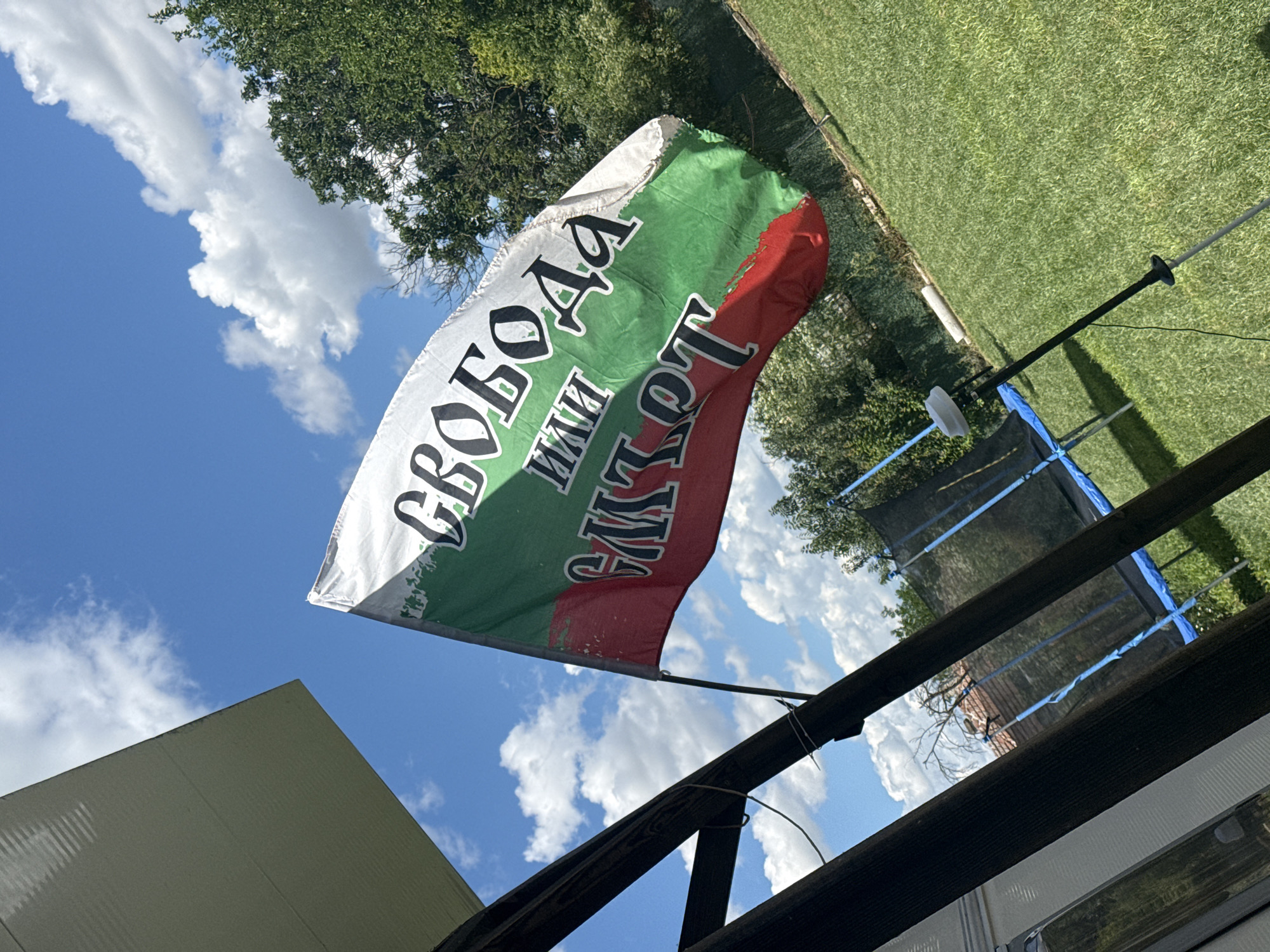 The image depicts a Bulgarian flag waving against a bright blue sky with scattered white clouds. The flag is horizontally striped with white, green, and red. Text in black lettering is visible on the white and green sections of the flag. The bottom portion of the flag shows a distressed red and green area. The flag is attached to a pole that is partially obscured by dark wooden beams, suggesting it is flying in a backyard or outdoor area. To the left, a portion of a white structure, possibly a roof or a wall, is visible. The background includes a lush green lawn, trees, and a trampoline. The scene is outdoors, under natural daylight. The specific location is identified as Rusokastro, Bulgaria.