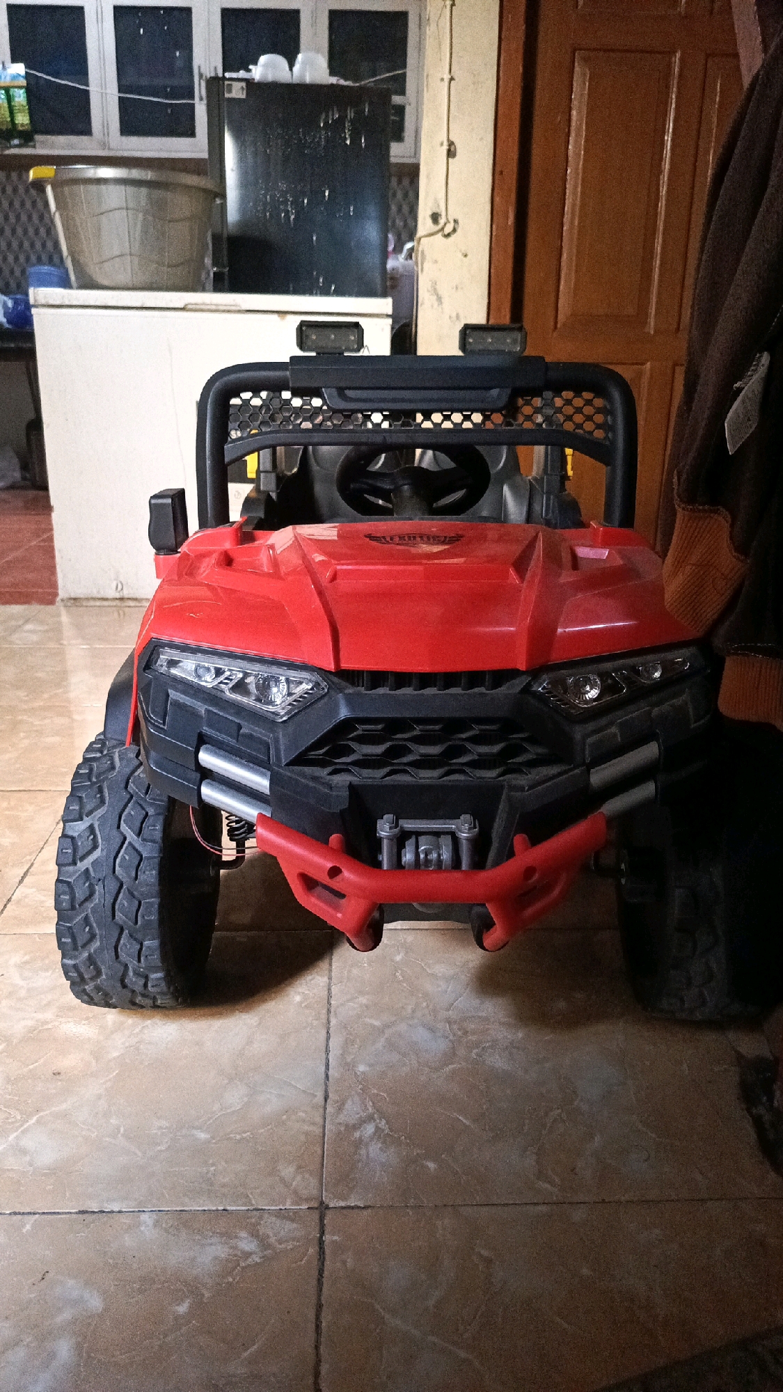 An indoor scene features a bright red toy ATV. The ATV has a black roll cage, headlights, and a grill. It is parked on a tiled floor in what appears to be a residential area. In the background, there is a black refrigerator, a white chest freezer, and a brown wooden door. The overall lighting suggests it might be evening or a dimly lit room. The location is identified as Pekauman Kulon, Indonesia.