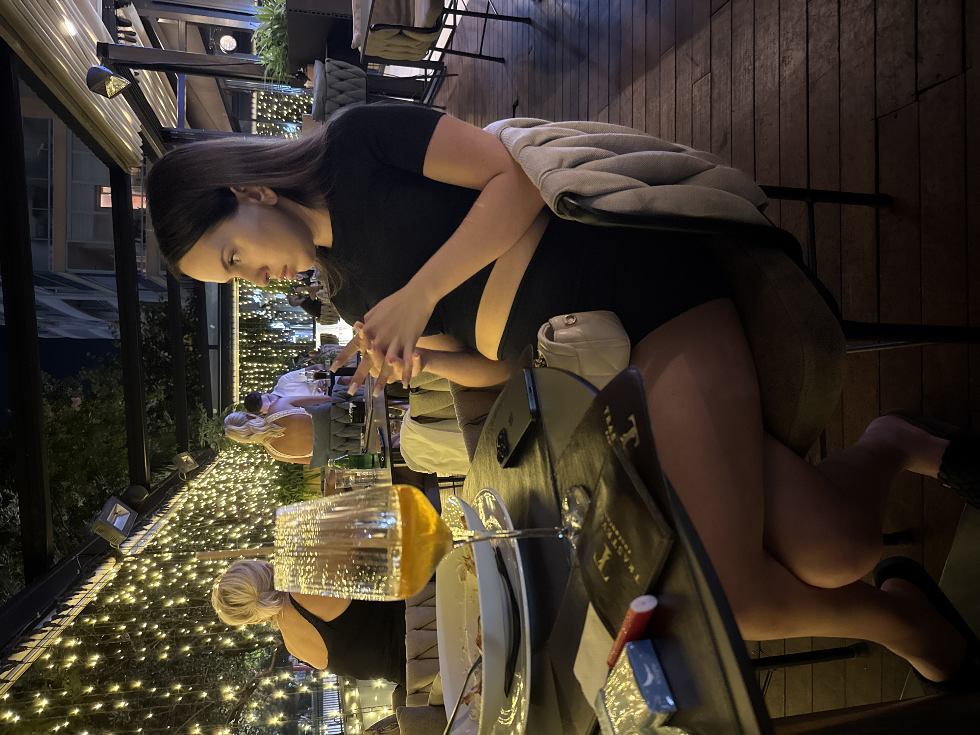 A woman with dark hair is seated at a table outdoors at night, her profile facing right. She is wearing a black crop top and black shorts. Her hands are clasped in front of her. A glass of amber liquid sits on the table, as do a pack of cigarettes, a red lipstick, and a smartphone. Another woman with blonde hair is seated at a table behind her, facing away from the camera. The background is decorated with strings of small, warm-toned lights, and there are plants visible. The flooring is made of wooden planks.