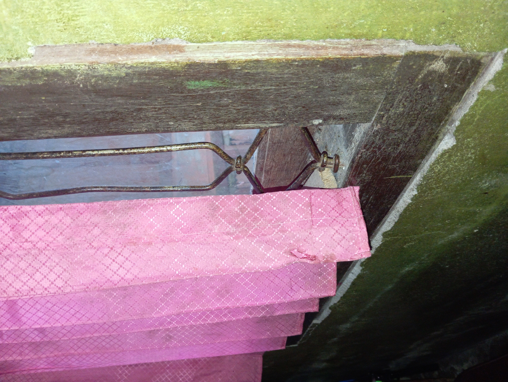 A close-up, eye-level, medium shot shows a section of an old, weathered wooden window frame and a tattered pink curtain. The window frame is dark brown, with visible wood grain and some green moss or algae growing on its surface. A rusty metal window guard with a decorative, angular design is visible behind the curtain. The curtain itself is a vibrant pink with a repeating diamond pattern. It is drawn partially closed, revealing several pleats. A noticeable tear is present in the lower right portion of the curtain. The wall surrounding the window frame is painted a light green color, with patches of lighter colored plaster. The scene appears to be an interior view from a room in Rejosari, Indonesia, likely showing a window in a residential building.