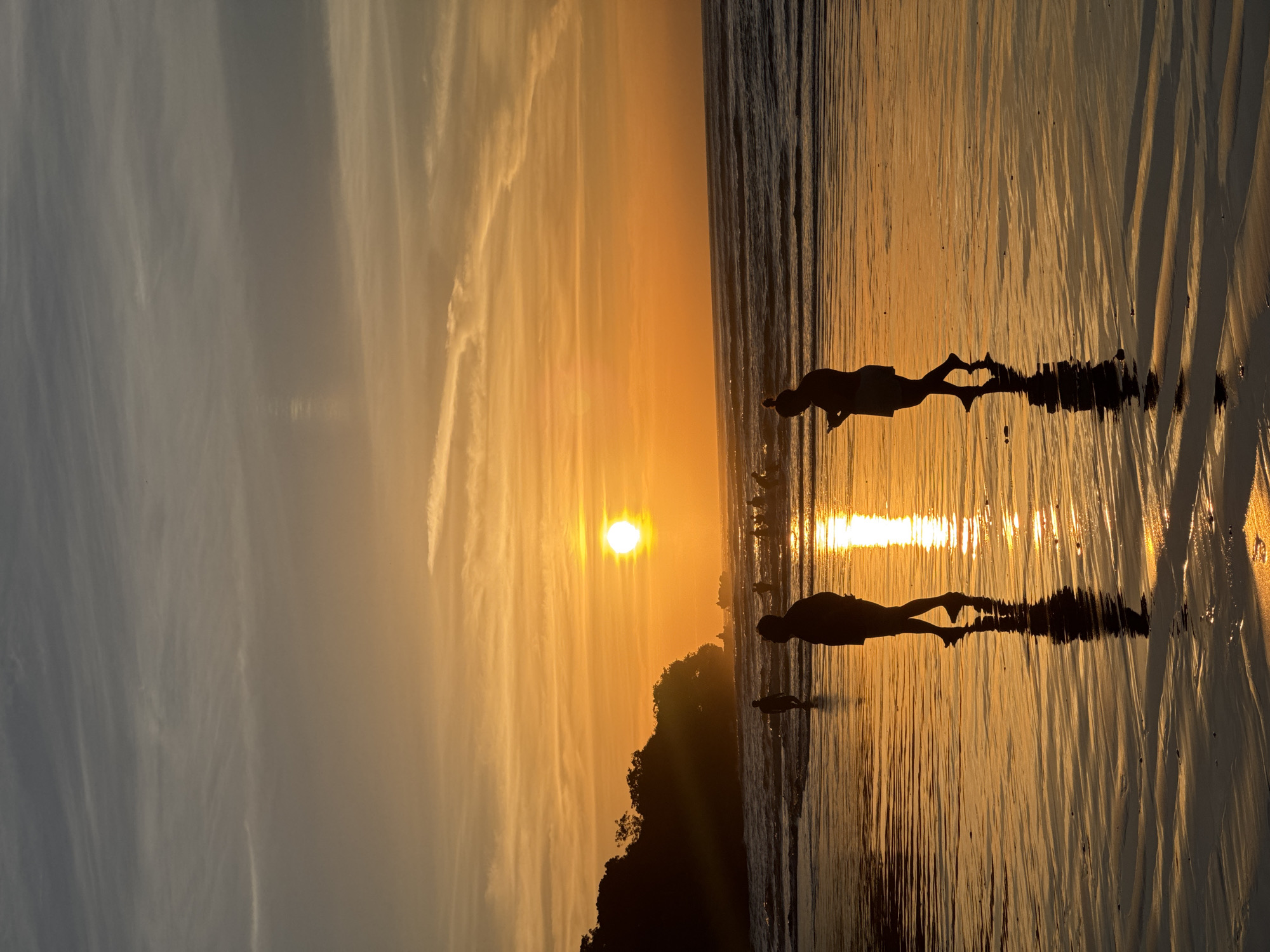 The image depicts a sunset scene at a beach in Kuta Selatan, Indonesia. Two silhouetted figures are visible wading in the shallow water, their reflections mirrored on the wet sand. The sun is positioned on the horizon, casting a bright golden light across the sky and water. A tree line is faintly visible in the mid-ground between the sun and the figures. The sky shows a mix of orange and gray hues with streaks of clouds. The wet sand reflects the sunlight, creating vertical patterns.