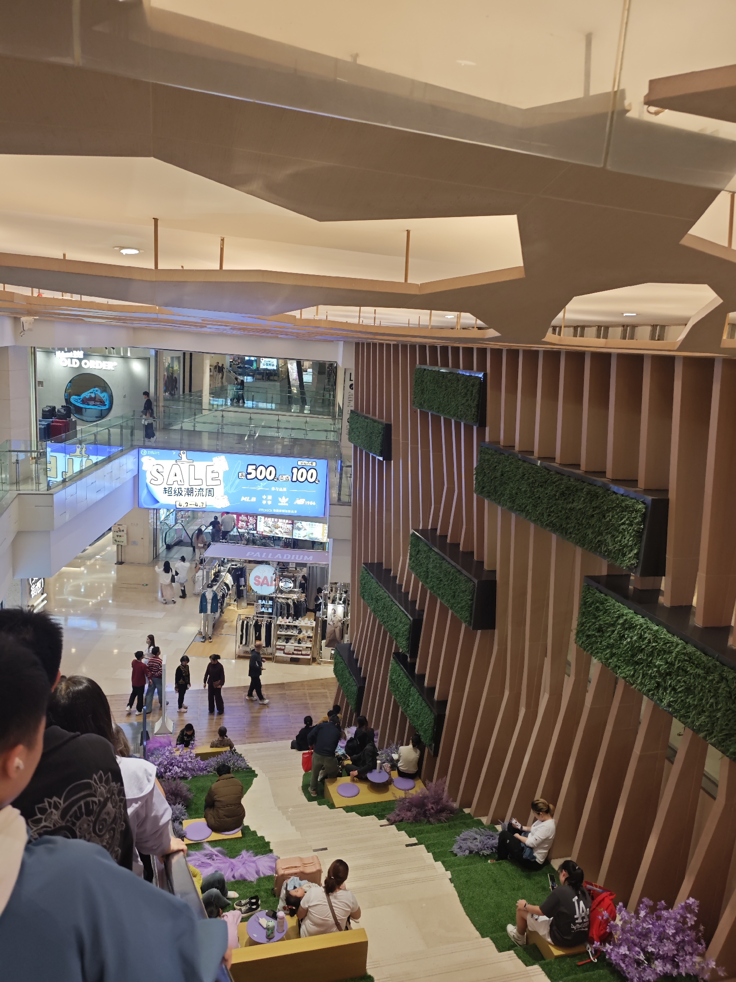 The image shows the interior of a shopping mall in the Yuzhong District of China. People are walking around different storefronts and levels. There is a "SALE" sign hanging above the first level. People are sitting on the steps and a grassy area on the right of the image. The background contains wooden panels with a green foliage motif. The ceiling features a modern design with wooden elements. Shoppers are seen browsing merchandise, sitting, and moving through the mall.