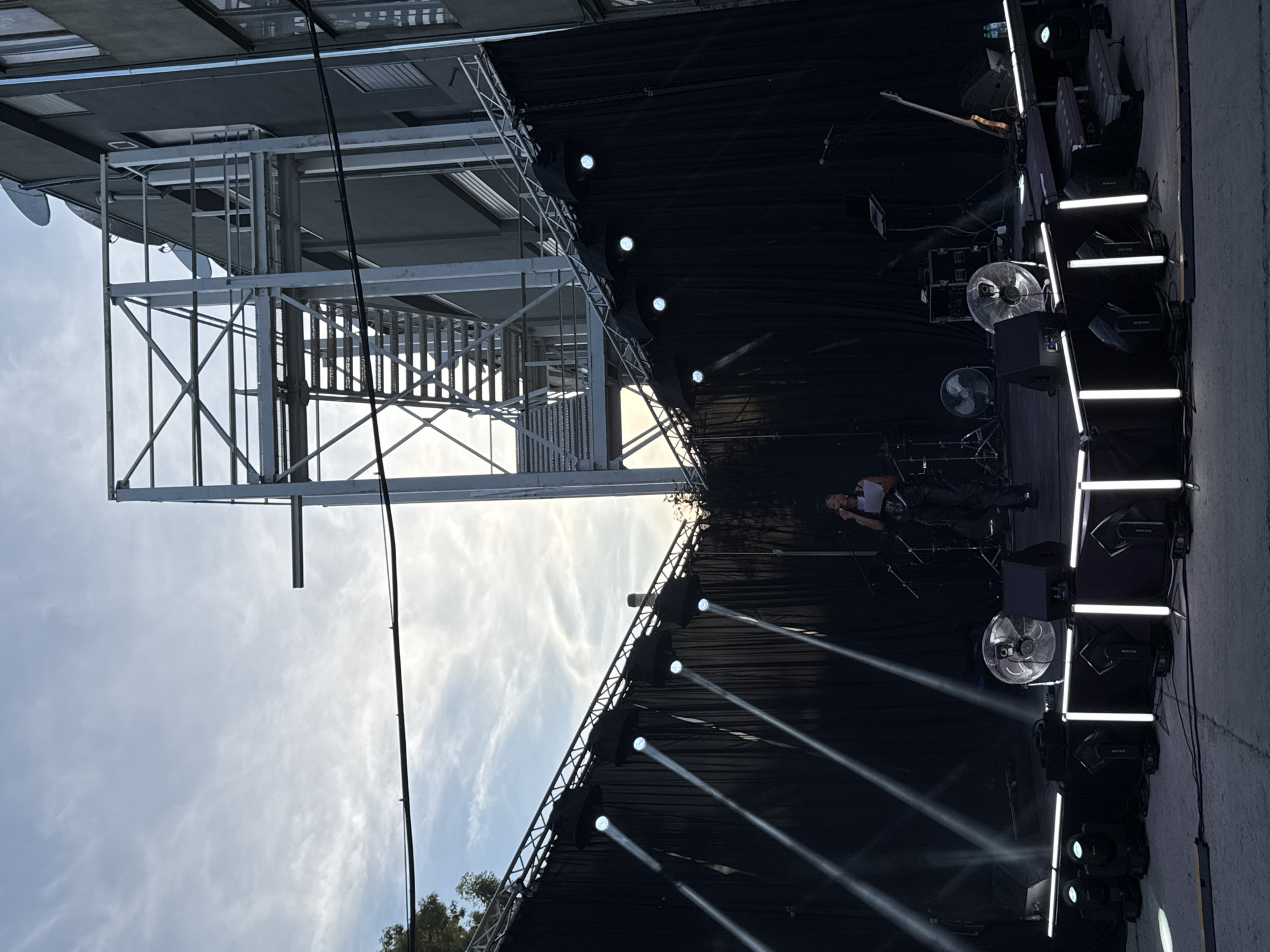 The image depicts an outdoor stage setup, likely for a performance. A woman is standing on the stage, holding a paper and speaking into a microphone. She is wearing black pants and a white top. Behind her, there are drums and other musical equipment. The stage is draped with black curtains and illuminated by stage lights, some of which are angled downwards, creating beams of light. In front of the stage, there are several lighting fixtures and fans. In the background, a building with a metal fire escape is visible against a cloudy sky. The scene suggests a prepared event, possibly a concert or festival, taking place in an urban or semi-urban environment. The presence of the fire escape and building facade indicates a location within a city, though no specific landmarks are identifiable that would confirm the location as Busmantsi, Bulgaria.