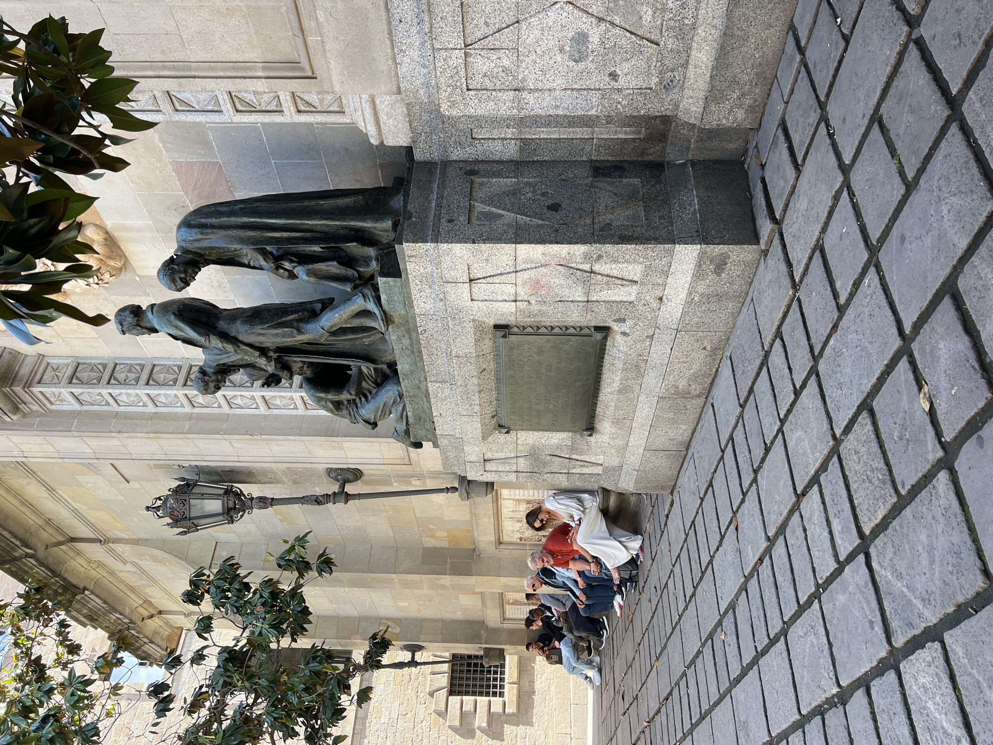 The image shows a street scene in Barcelona, Spain, featuring the Monument a Bartomeu Robert. The monument is made of bronze and stone and features a statue group. A group of people is sitting on the edge of the monument's base. The ground is paved with cobblestones. A decorative lamppost is visible, along with some greenery from trees.