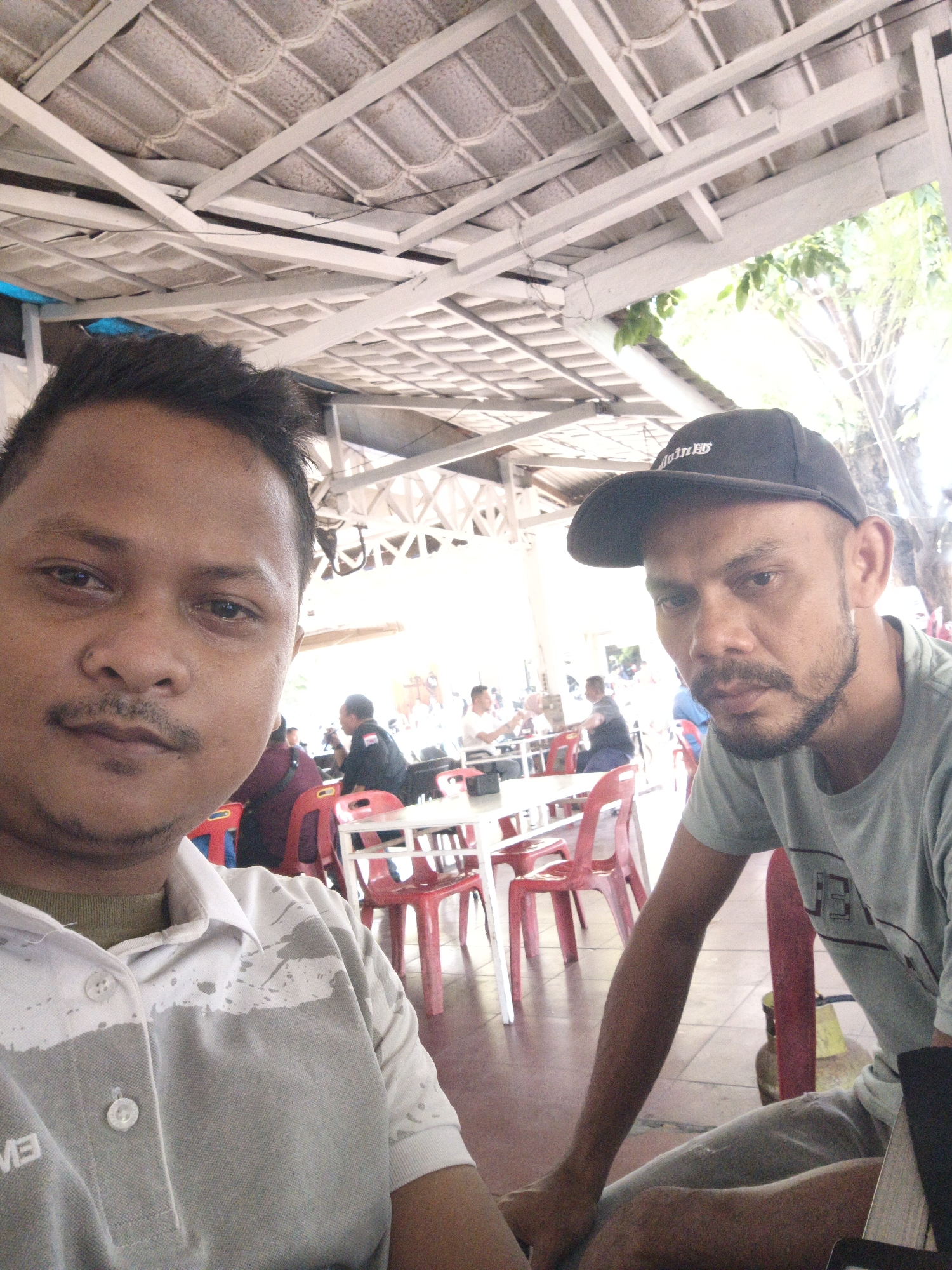This image depicts a semi-outdoor dining or gathering area located in Lhokseumawe, Indonesia, characterized by strong natural daylight.
In the foreground, two adult males are visible, both looking towards the camera. The individual on the left has short dark hair, a moustache, and a short beard. He wears a collared, short-sleeved polo shirt, grey on the lower torso, transitioning to a white upper section with irregular grey patterns. An olive-green crew-neck shirt is visible beneath the polo. The individual on the right has short dark hair, a beard, and a moustache, and wears a black baseball cap with partially visible white text "JOINED" on the front. He is dressed in a light olive-green short-sleeved t-shirt. He appears to be seated or leaning forward, positioned slightly behind and to the right of the first man.
The environment features an overhead roof structure composed of light-colored corrugated tiles, possibly asbestos cement, supported by a framework of white-painted beams and trusses. Below, the floor is covered with reddish-brown square tiles. Multiple red plastic chairs are arranged around white rectangular tables in the midground and background, where several other individuals are seated. Their actions suggest typical social engagement in a dining setting, with at least one person in the midground wearing a dark shirt with a patch on the left sleeve. Green foliage and tree trunks are visible beyond the covered area on the right side of the image. A yellow object, possibly a gas cylinder, is partially visible on the floor in the lower right background.