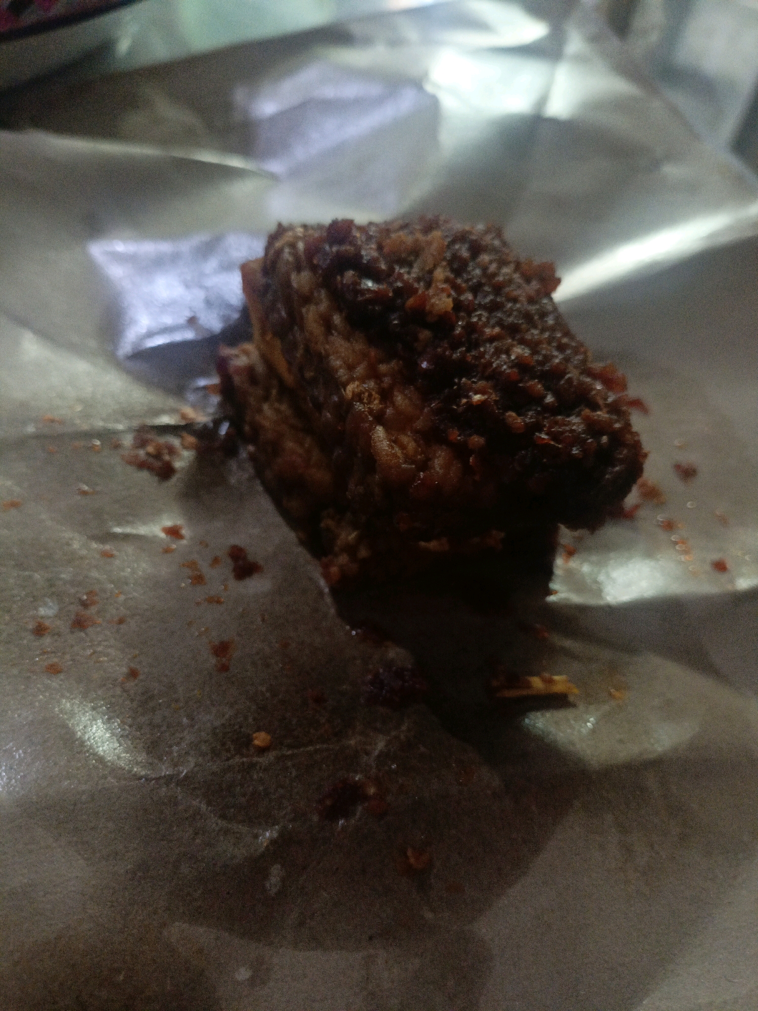 The image shows a close-up of a piece of fried food, possibly chicken or meat, with a dark, craggy exterior, resting on a crumpled piece of paper. The paper appears to be textured and slightly reflective, with small reddish-brown crumbs scattered across its surface, particularly around the food item. The lighting is dim, creating shadows and highlighting the texture of the food and paper. The background is out of focus and appears to be a metallic or reflective surface, possibly aluminum foil. The overall impression is of a casual, perhaps street food, setting. The specific location in Undaan, Indonesia, cannot be determined from the image alone, as it lacks any discernible landmarks or contextual clues.