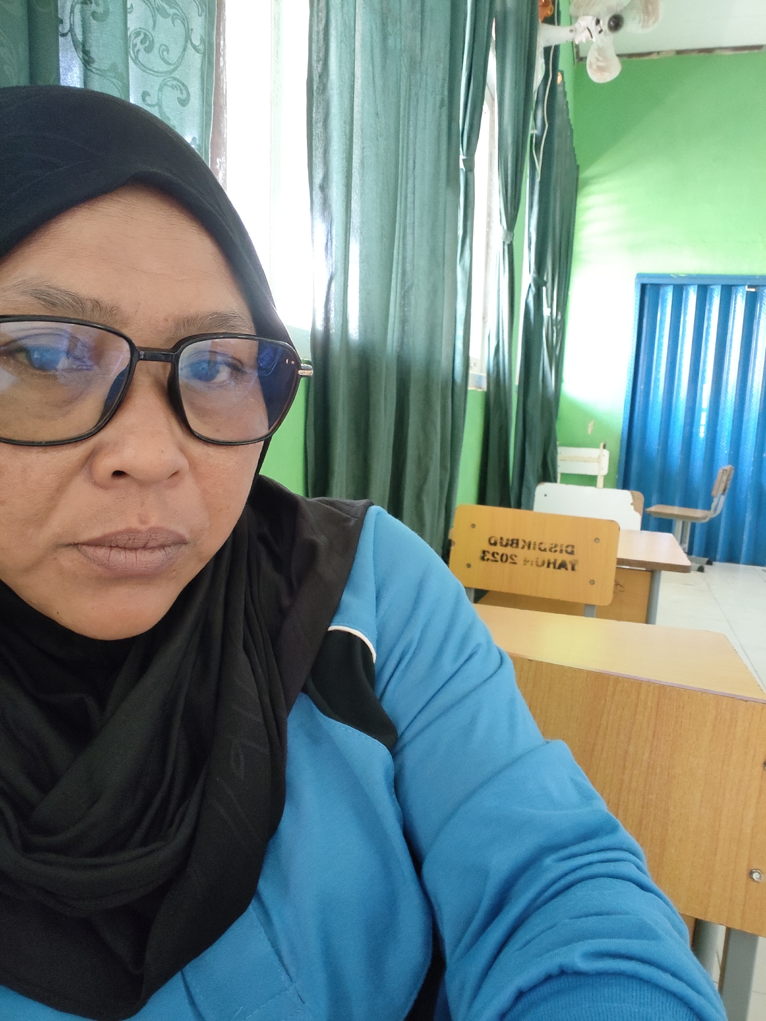 An Indonesian woman, wearing glasses and a black hijab, is seated at a desk in a classroom. She is wearing a blue shirt with black and white accents. Behind her, green curtains are visible, as well as a white ceiling fan. The classroom also contains wooden desks and chairs, and blue corrugated doors. The image appears to have been taken in Lori, Indonesia.