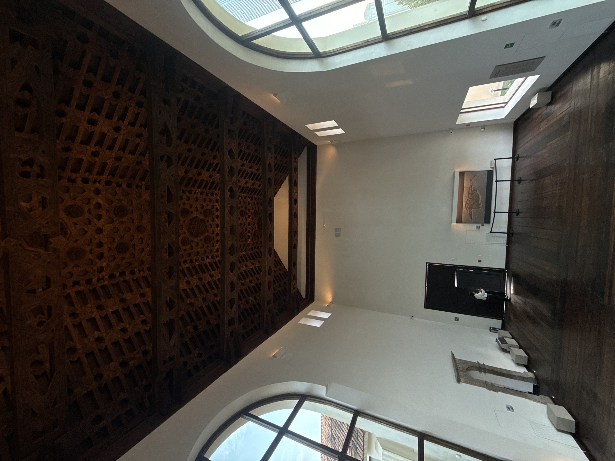 The image depicts an interior view of the Museo El Centenario in San Pedro Garza García, Mexico. The architecture showcases a blend of traditional and modern elements. A heavily ornamented wooden ceiling with intricate geometric patterns dominates the left side of the frame. The ceiling transitions into a white wall with skylights and windows, allowing natural light to illuminate the space. The right side of the frame features a dark wooden wall. Architectural fragments and a black door with figures inside are present on the right.