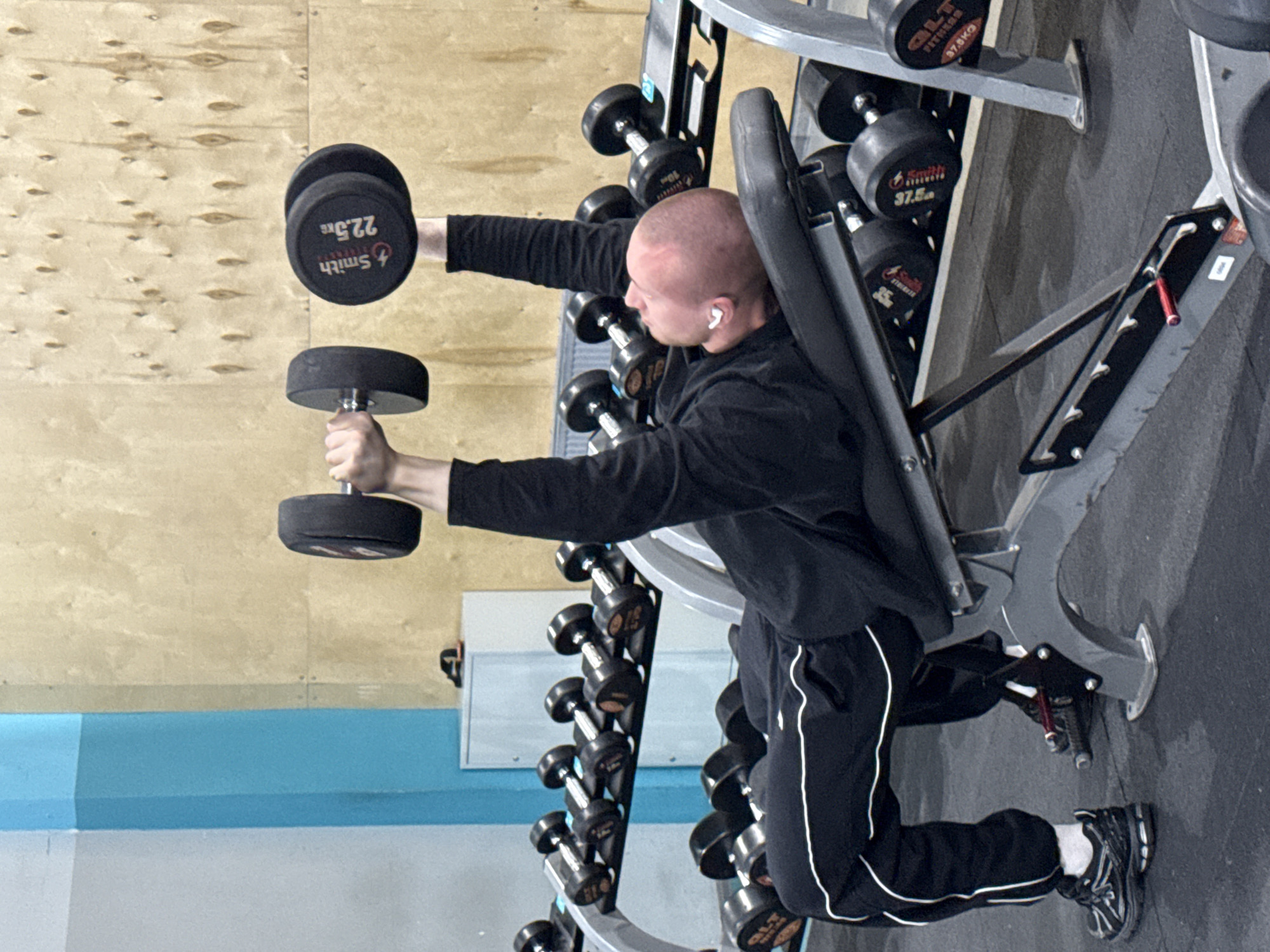 A man is exercising with dumbbells in a gym. He is positioned next to a weight rack with various dumbbells. He's wearing a black long-sleeved shirt and black sweatpants with a white stripe. He has white earbuds in his ears. The dumbbells in his hands are black, one weighing 22.5kg and the other obscured. The gym's walls are primarily light brown wood paneling with a light blue accent stripe. The weight rack contains a variety of dumbbells labeled "Smith Fitness" and "QLT Fitness", with weights ranging from 10kg to 37.5kg. The location appears to be a gym somewhere, it is impossible to determine if it is located in the city of Yekaterinburg, Russia or anywhere else in the world.