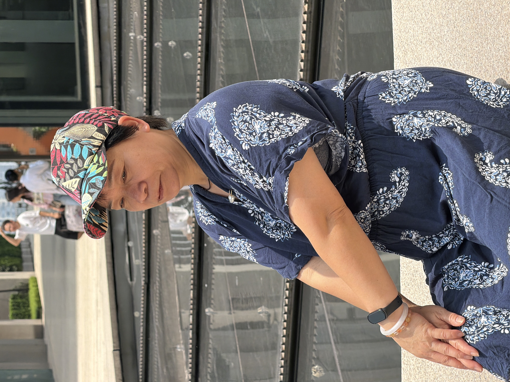 A medium shot shows an East Asian woman sitting outdoors in what appears to be an urban park or plaza in Yuzhong District, China. She is wearing a dark blue, short-sleeved top with a white paisley pattern, a matching skirt, and a colorful, patterned baseball cap. She is looking to her left with a neutral expression. Her left arm is bent, resting on her lap, and she has a black smartwatch and two bracelets on her left wrist. In the background, there are reflections on tiered structures, possibly water features or glass panels. Two other people, one man in a white t-shirt and shorts and a woman in a light pink top, are visible in the background, walking away from the camera. The ground is a light tan color, and there are buildings and greenery in the distance. The lighting suggests a sunny day.