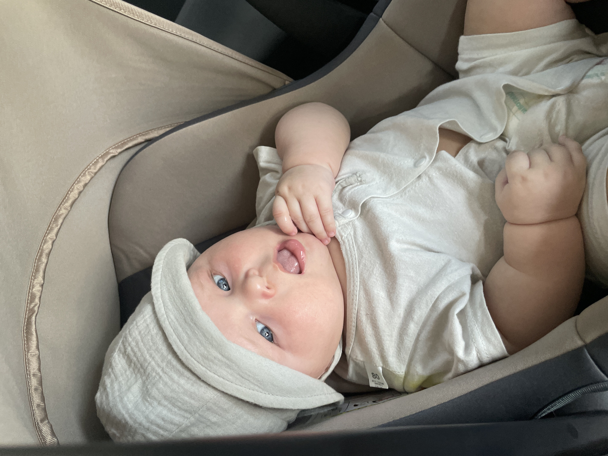 A baby wearing a light-colored hat and onesie is in a stroller. The baby has bright blue eyes and is looking upwards with its mouth slightly open. The stroller appears to be inside a vehicle, with the interior visible in the background. There are no visible indications of the city of Stavropol, Russia.