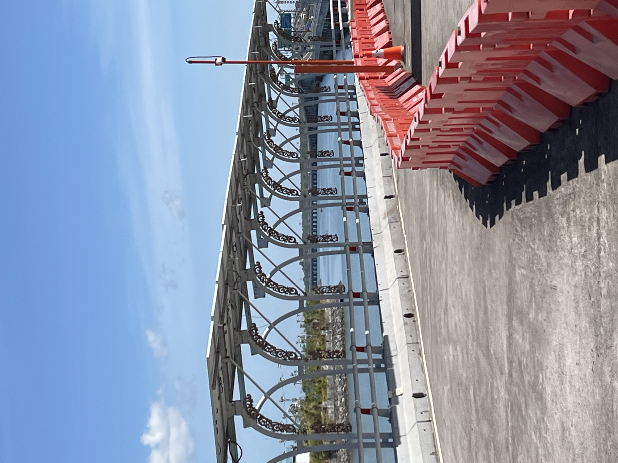 The image shows a bright, sunny day with a clear blue sky and scattered clouds. In the foreground, a grey asphalt road curves to the right. A bright orange plastic barrier with red and white reflective stripes runs along the right side of the road, creating a temporary traffic management system. A traffic cone is visible behind the barrier.
In the mid-ground, a pedestrian walkway or bridge is visible. It is covered by a structure with solar panels on top, supported by metal beams and decorative arches. The walkway has a grey concrete barrier along its edge, with circular openings visible at intervals. Beyond the walkway, a body of water is visible, with a bridge extending across it in the distance. Lush green vegetation and trees can be seen on the shore to the left of the walkway.
The scene is likely the **Benoa Bridge** in South Denpasar, Bali, Indonesia, which is known for its distinctive solar panel-covered walkway and its location over a bay. The presence of construction barriers suggests ongoing work or maintenance. No people are visible in the image. The overall impression is one of a modern infrastructure project in a coastal environment.