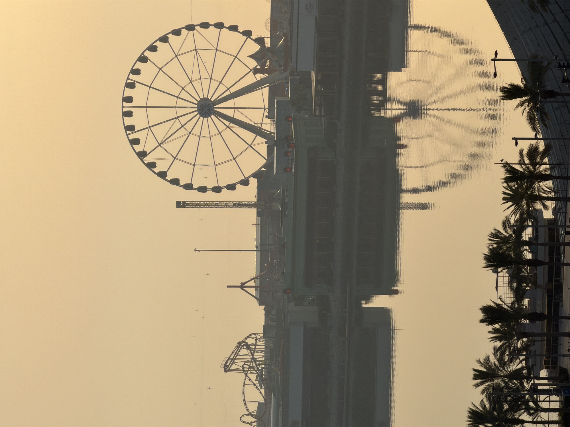 The image depicts an amusement park with a prominent Ferris wheel and a roller coaster, silhouetted against a hazy, light orange sky. The structures are reflected in the calm water in the foreground. Palm trees are visible along the edge of the water, and some buildings and lights are present in the background. The scene appears to be at dusk or dawn due to the soft, diffused light.