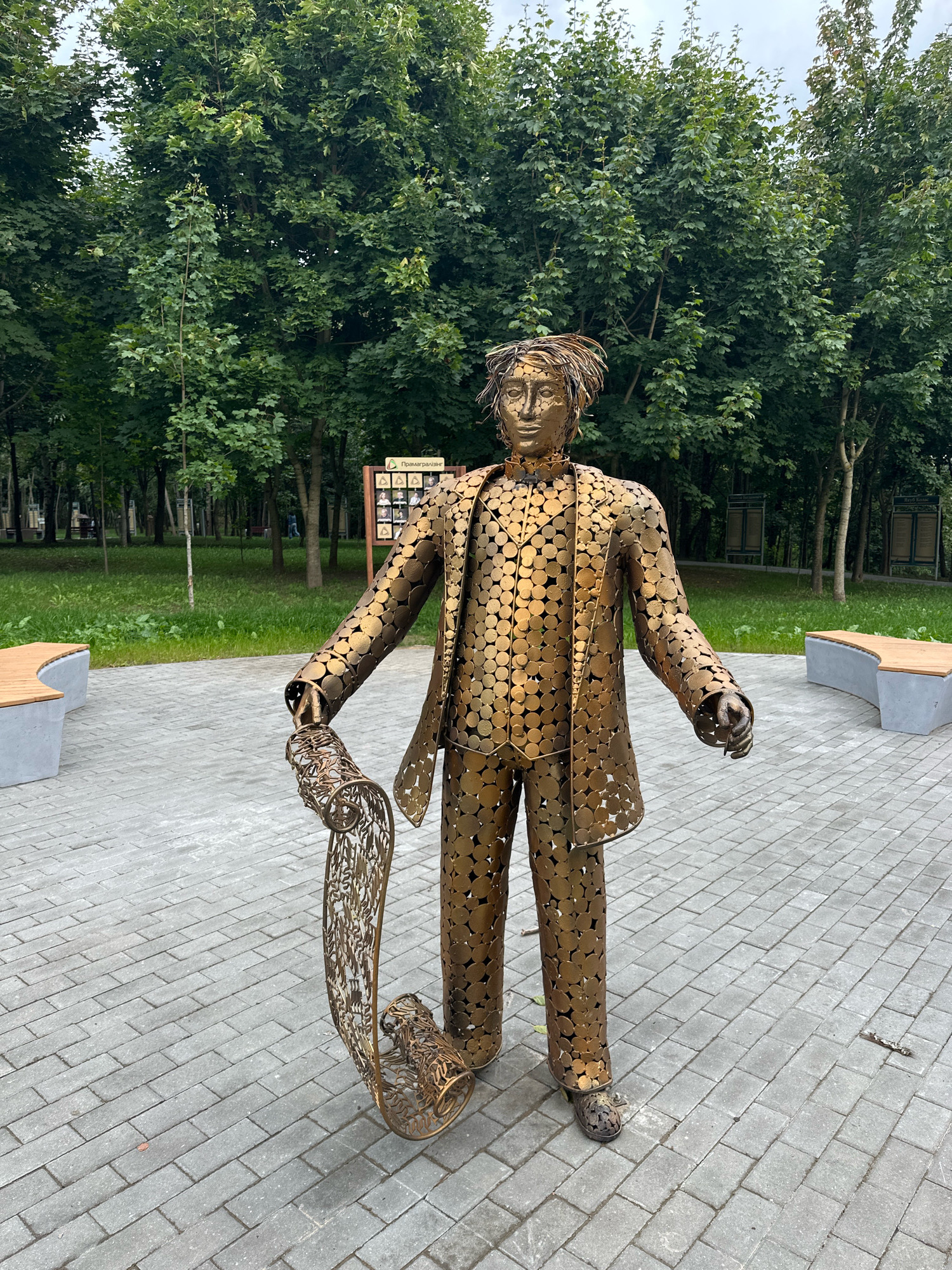 A full-body, outdoor, eye-level shot shows a metallic sculpture of a man in a suit, holding a scroll, located in a park in Minsk, Belarus. The sculpture is made of numerous golden, circular metal pieces, giving it a textured appearance. The man has a slightly disheveled hairstyle and is facing forward with his arms outstretched. He is holding a large, ornate scroll in his left hand. The sculpture stands on a paved pathway made of grey rectangular bricks.
In the background, there are several lush green trees, typical of a park setting. To the left and right of the sculpture, there are curved park benches with wooden seats and grey concrete bases. In the middle ground, behind the sculpture and to the right, there is a wooden information board with multiple framed pictures. The ground is covered with grass. The sky is overcast.