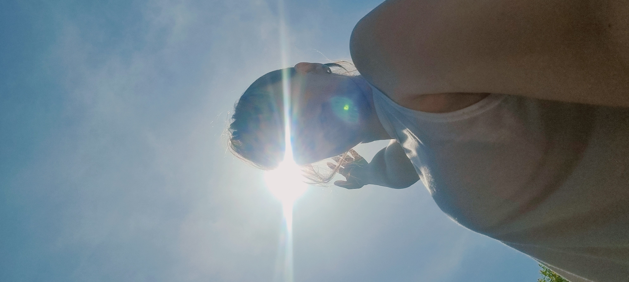 An upward-angled outdoor shot under bright sunlight features a person looking up at the sun, their head partially obscuring the sun itself. The sky is clear blue. The person has fair skin and is wearing a white top. Some vegetation is faintly visible in the bottom right corner. The location is Dunnae-myeon, South Korea.