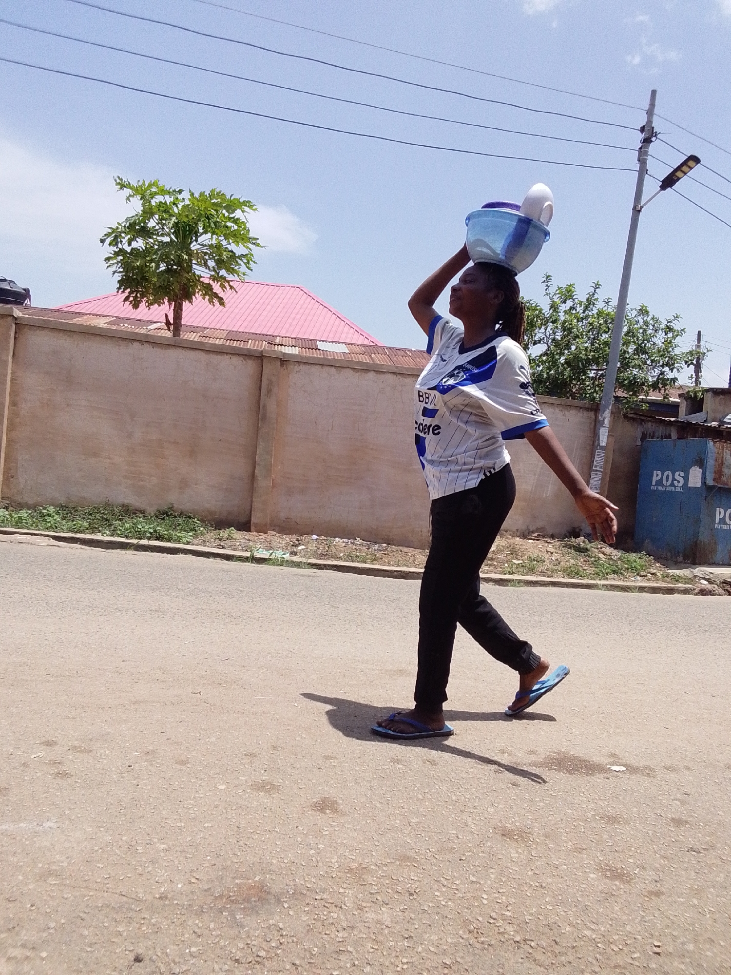 A person wearing a white and blue jersey, black pants, and blue flip-flops is walking down a paved road. The person has a blue bowl and white plates balanced on their head. In the background, there is a beige wall with a green shrub at its base, and a building with a pink roof is visible behind the wall. A small green tree is growing just above the wall. A utility pole with a streetlight attached is to the right of the person. A small shed with "POS" printed on it is partially visible to the right. The sky is blue with a few white clouds.