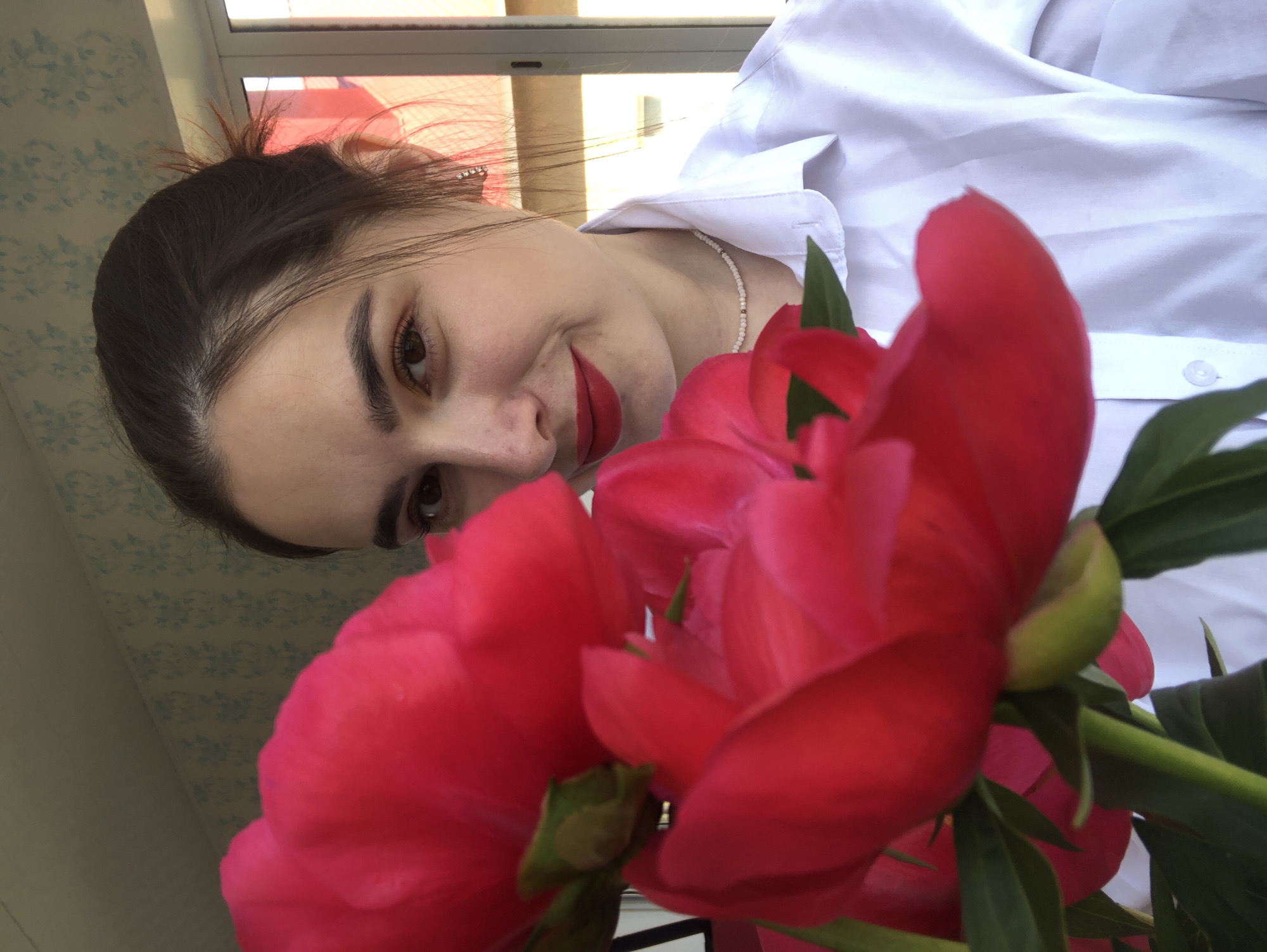 The image depicts a close-up, eye-level shot of a young woman of Caucasian descent, positioned in the foreground to the right, holding a bouquet of bright pink peonies. She has dark brown hair pulled back loosely and is wearing a white collared shirt and a delicate beaded necklace. Her makeup includes bold red lipstick and defined eyebrows. The peonies, a dominant feature, are vibrant and take up a significant portion of the left side of the frame, their petals slightly out of focus.
The background reveals a wall with light blue floral wallpaper and a window with vertical blinds, suggesting an indoor setting. The lighting appears natural, possibly from the nearby window, casting soft shadows. The overall impression is one of a candid portrait with a focus on the subject and the flowers. The location is identified as Noviy, Russia, based on the provided information, but no specific landmarks are visible to confirm this within the image itself.