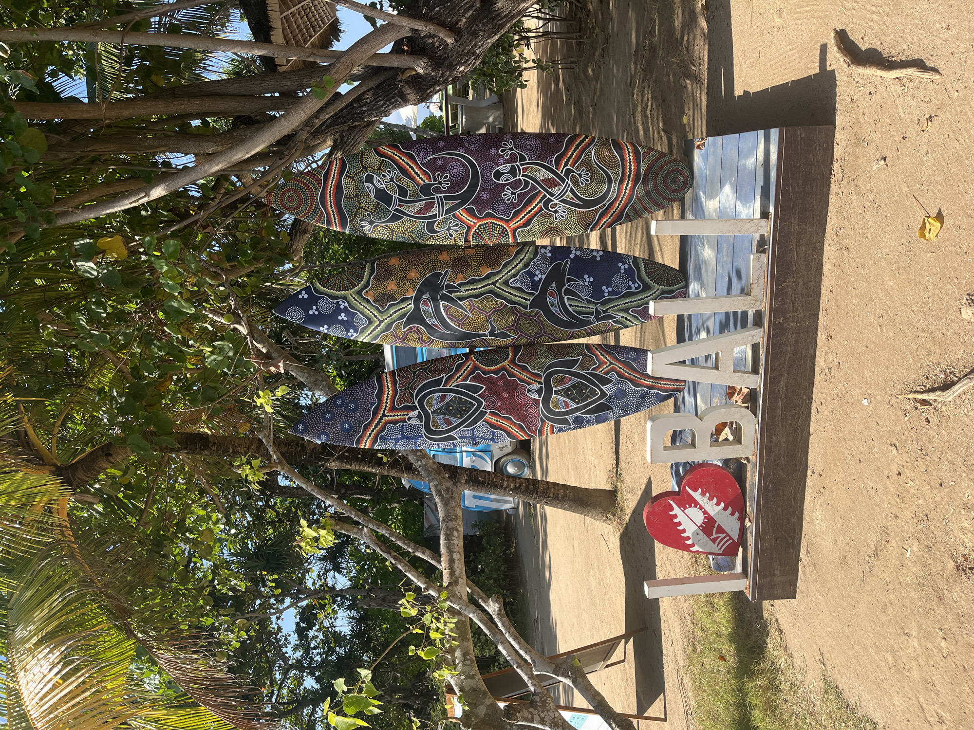 The image depicts an outdoor scene on a sandy beach, likely in Kuta Selatan, Indonesia, given the presence of surfboards and a tropical environment. In the foreground, a wooden sign spells out "I LOVE BALI" with a stylized red heart serving as the "O". Three surfboards with intricate Aboriginal-inspired art are propped up against a tree. These surfboards feature designs with turtles and lizards, rendered in a vibrant palette of blues, reds, yellows, and purples, with dot patterns. The surfboards are positioned in front of a light blue van, partially visible in the background, suggesting a relaxed beachside setting. Overhead, lush green palm trees and other tropical foliage provide shade, with sunlight dappling through the leaves. The ground is covered in sand and some scattered dry leaves. A few beach chairs and umbrellas are visible in the distance, hinting at a tourist-oriented location. The overall atmosphere is bright and sunny, characteristic of a tropical beach destination.