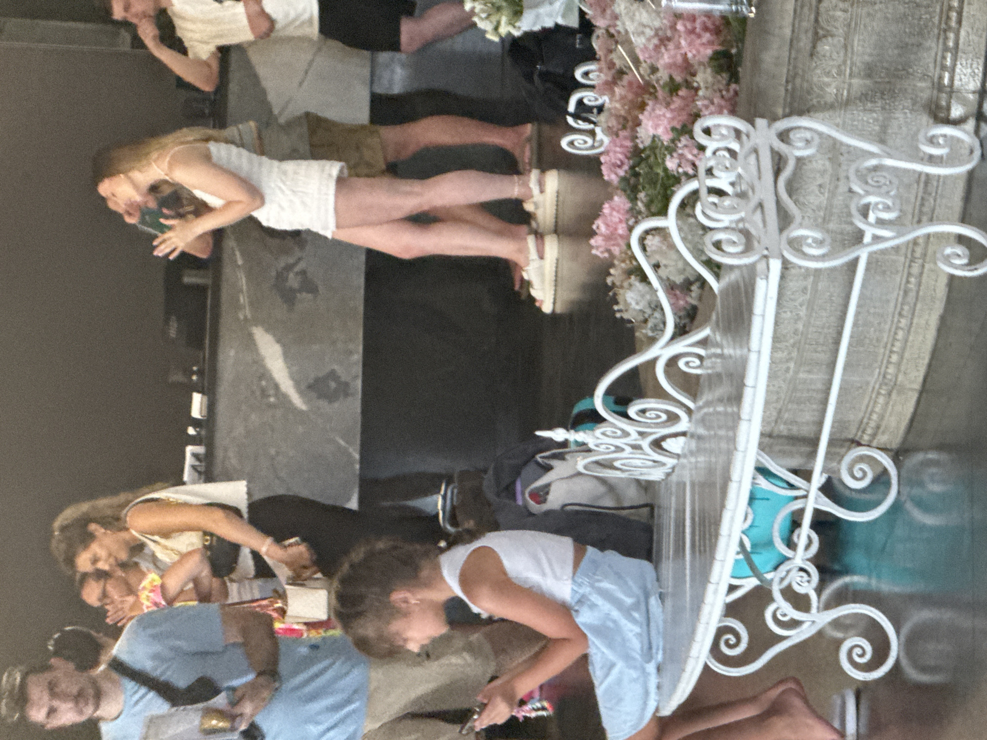 A group of people are gathered in what appears to be a public indoor space, possibly a hotel lobby or a venue in Yalta, Ukraine, near a large, dark, reflective counter. In the foreground, a young girl with dark hair is seated on a white bench, looking down at her phone. She is wearing a white sleeveless crop top and a light blue skirt. To her right, the bench is decorated with ornate white metalwork and is partially covered with floral arrangements in shades of pink and white. Behind her, a man in a blue t-shirt and khaki shorts stands holding a brochure. To the left of the counter, a woman with blonde hair in a ponytail is talking on her phone, wearing a white top and black pants. Further back, another woman in a white dress and shorts is seen from the side, also appearing to be engaged in conversation. Several other individuals are visible in the background, some standing and others walking, with bags and luggage suggesting travel. The overall environment appears to be modern and well-lit.