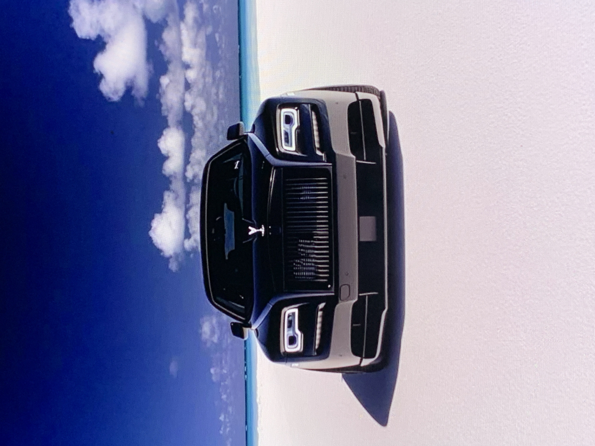 A black Rolls-Royce Phantom sedan is parked on a pristine white sand beach with the ocean and a bright blue sky in the background. The car is facing the camera and its headlights are on. The ocean is a vibrant turquoise and the sky is a deep blue with scattered white clouds. The scene is brightly lit, suggesting it is daytime.